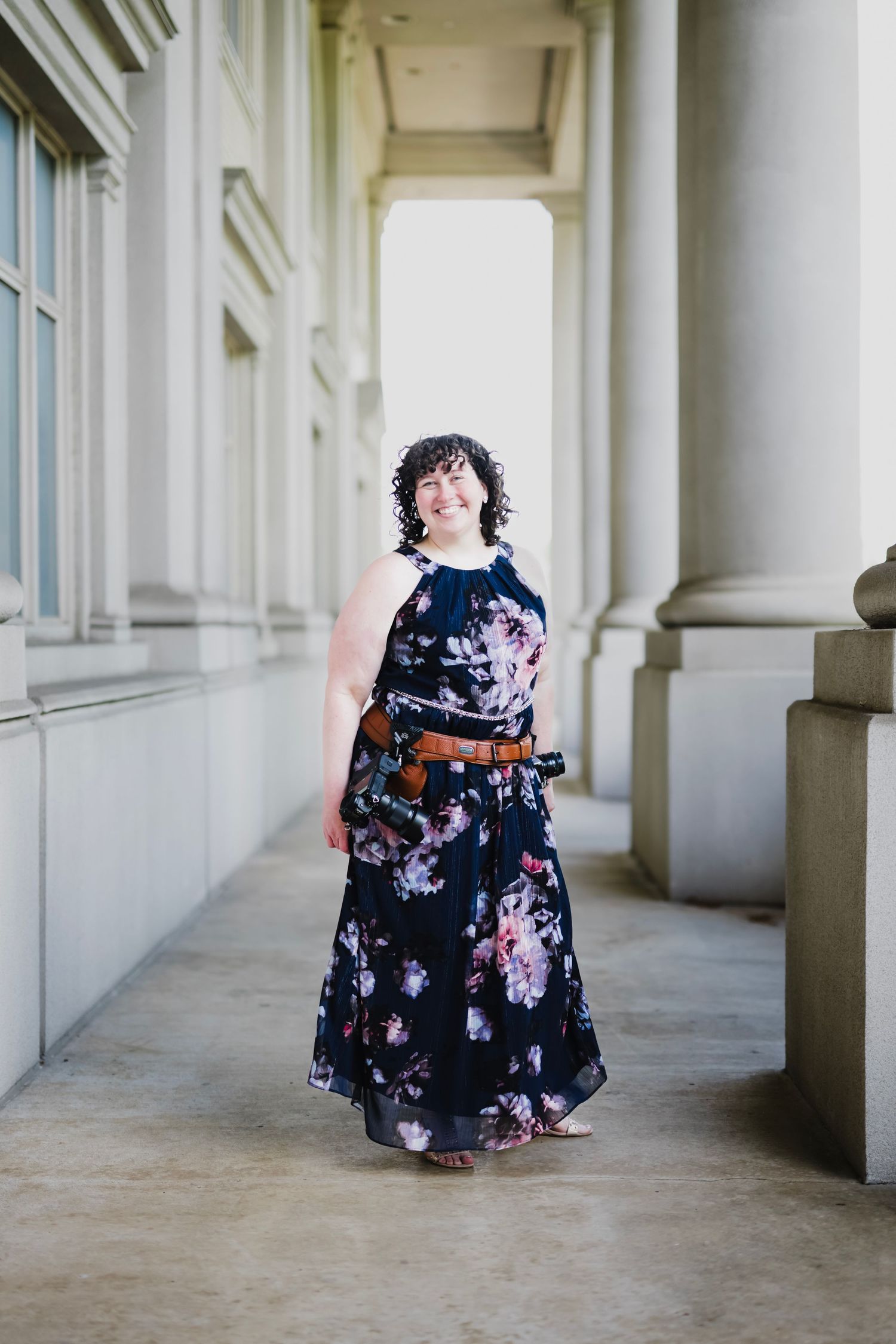 Woman in floral navy maxi dress with brown belt smiling between elegant white columns.