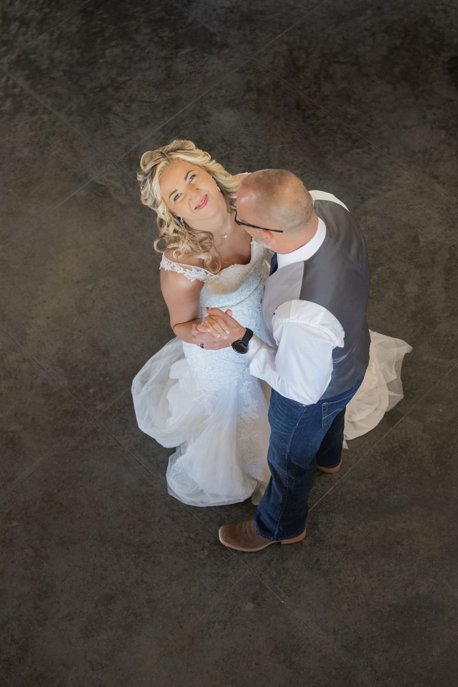 Aerial view of a bride in white gown and groom in jeans sharing a romantic first dance, looking up at camera.