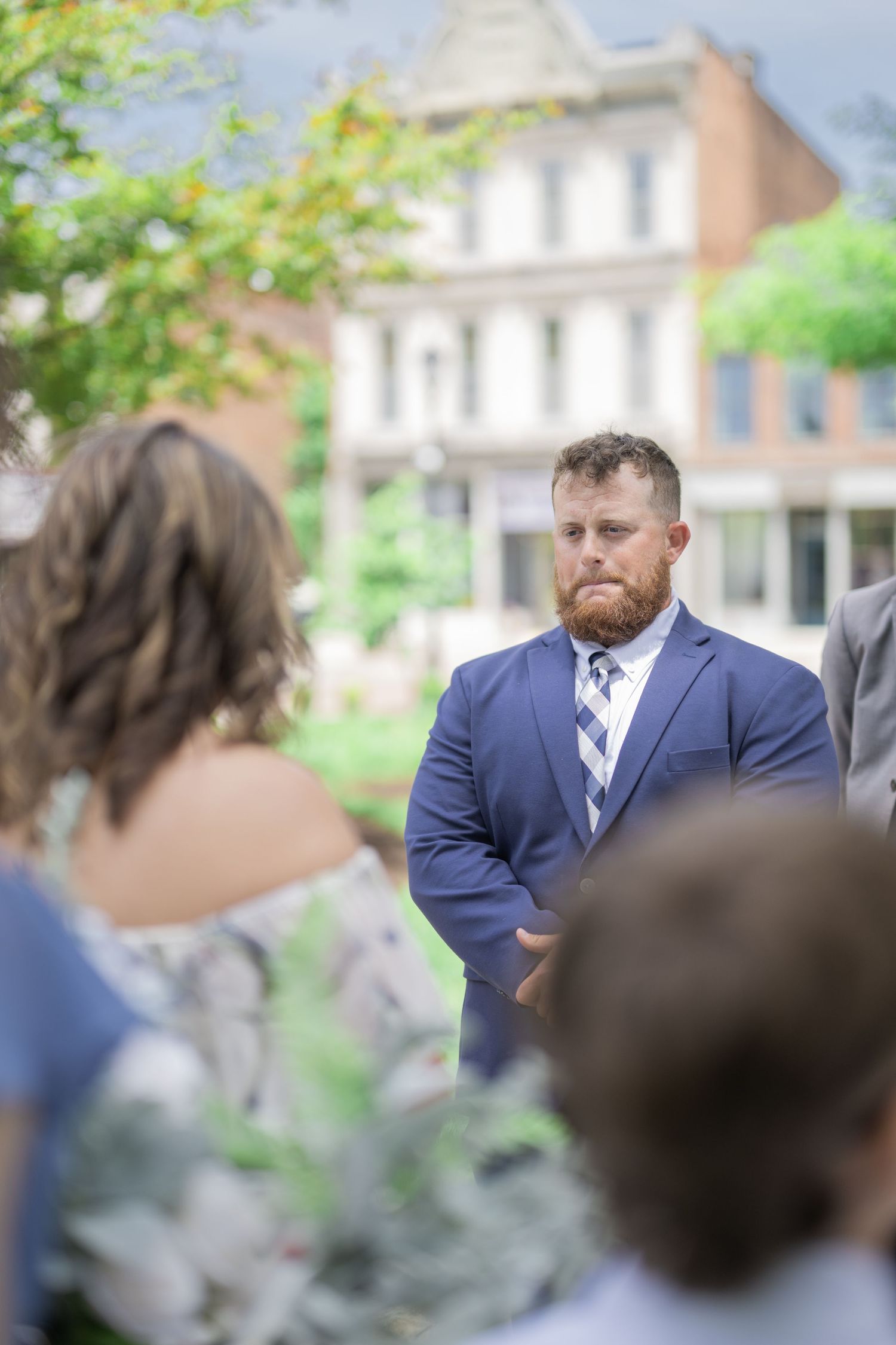 Groom in navy suit and plaid tie watches bride attentively during outdoor wedding ceremony.