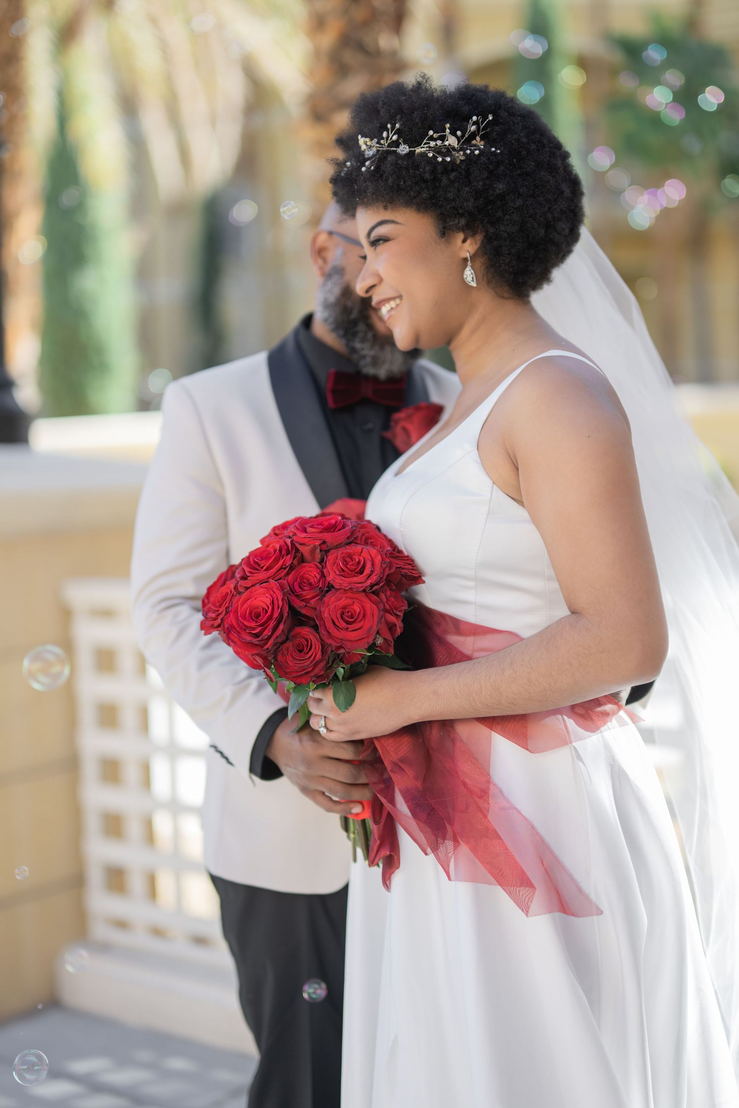 Smiling bride holding red roses bouquet in white gown with veil, groom in white tuxedo jacket standing behind her outdoors.