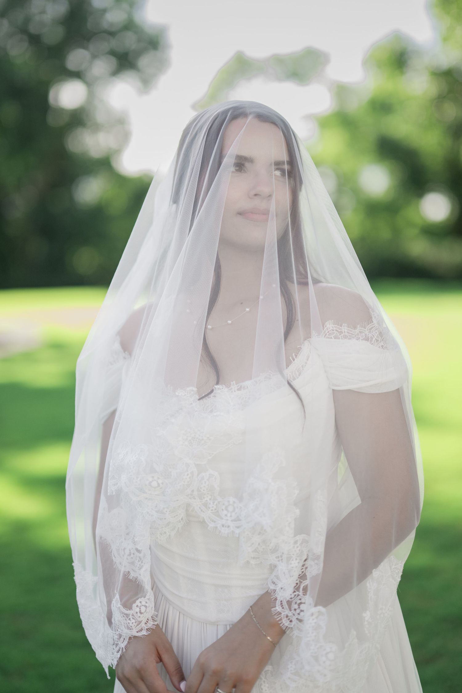 Elegant bride in white lace gown and veil standing outdoors in a lush green park setting.