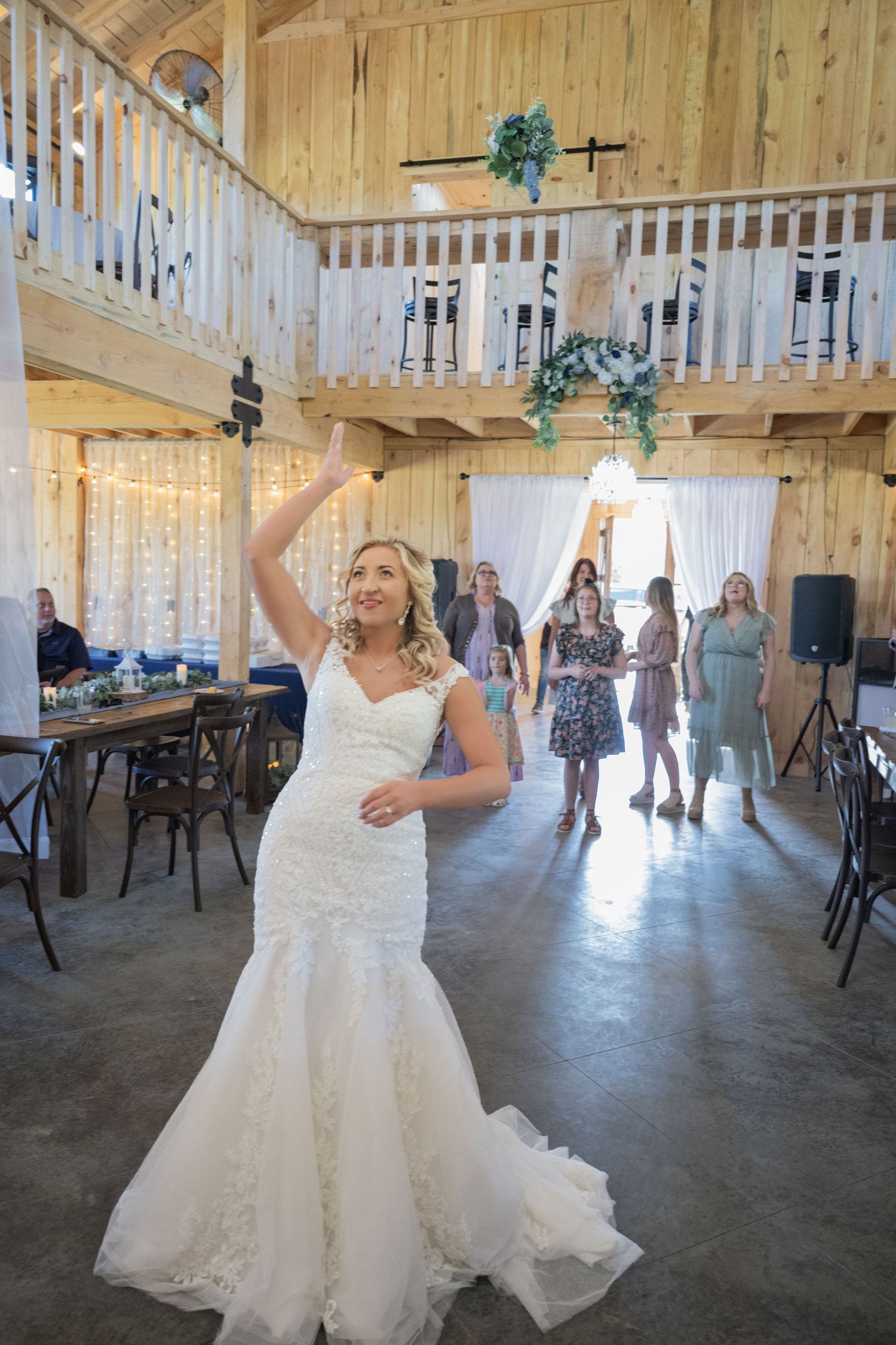 Bride in lace mermaid gown tosses bouquet to guests in rustic barn wedding venue with string lights.