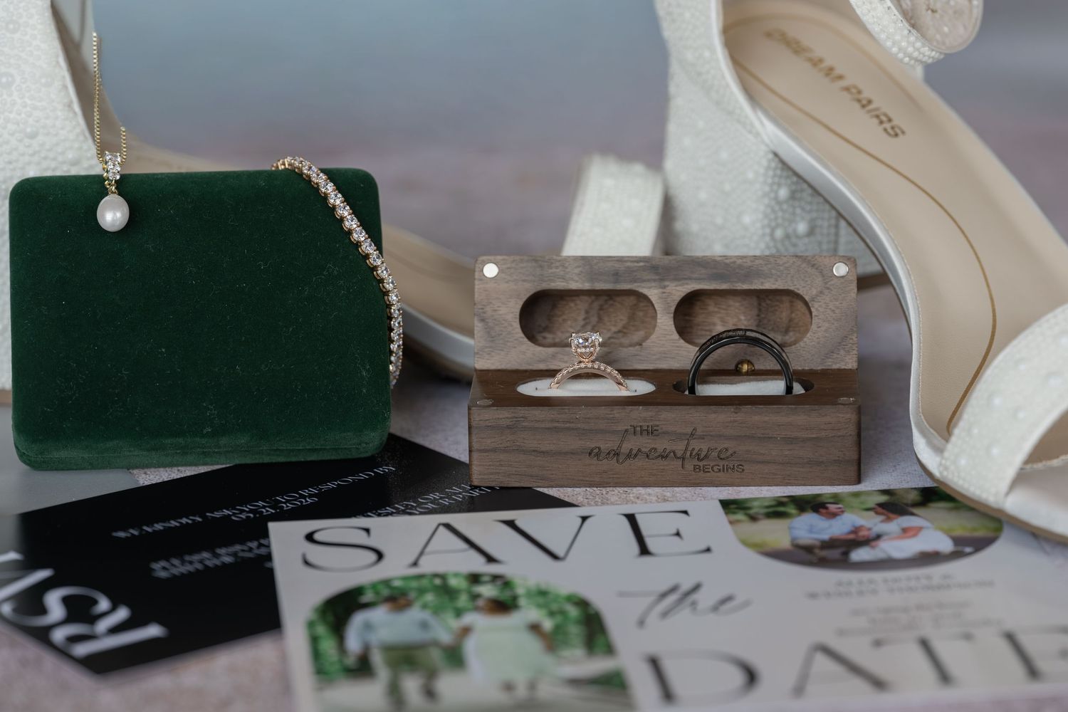 Wedding flat lay with rings in wooden box, green velvet jewelry holder, pearl necklace, heels, and save the date cards.