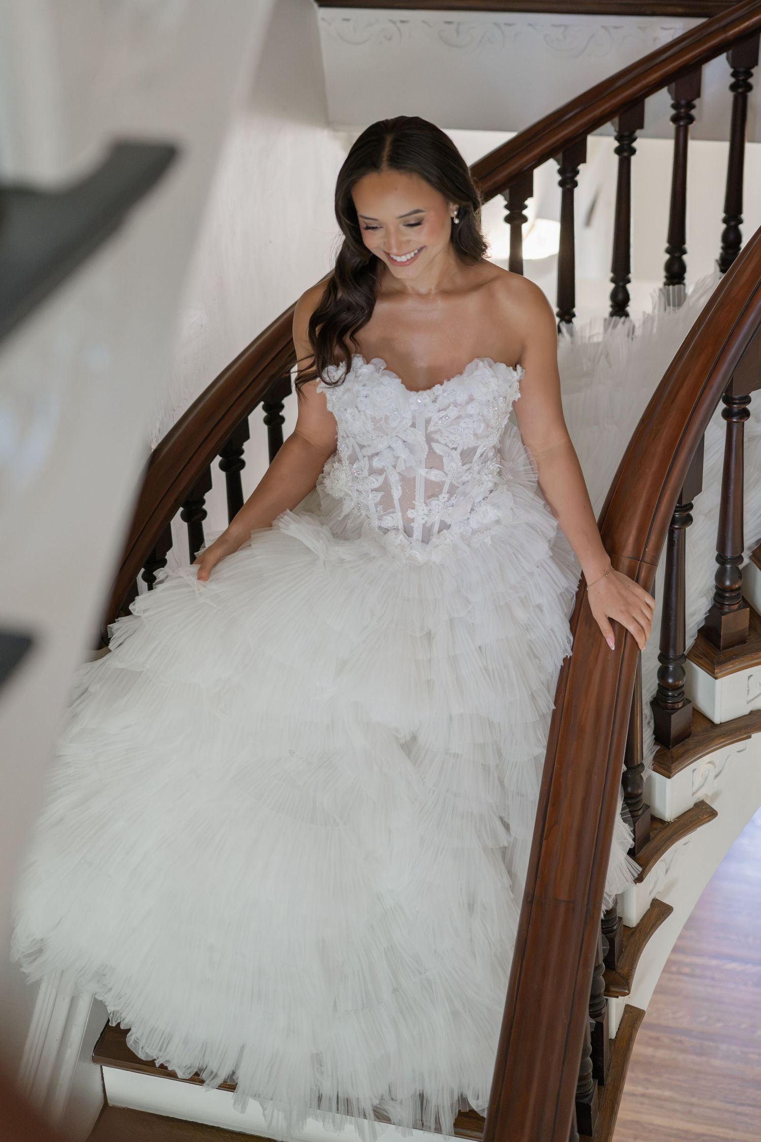 Bride in a strapless white ruffled tulle wedding gown descends an elegant curved wooden staircase, smiling gracefully.