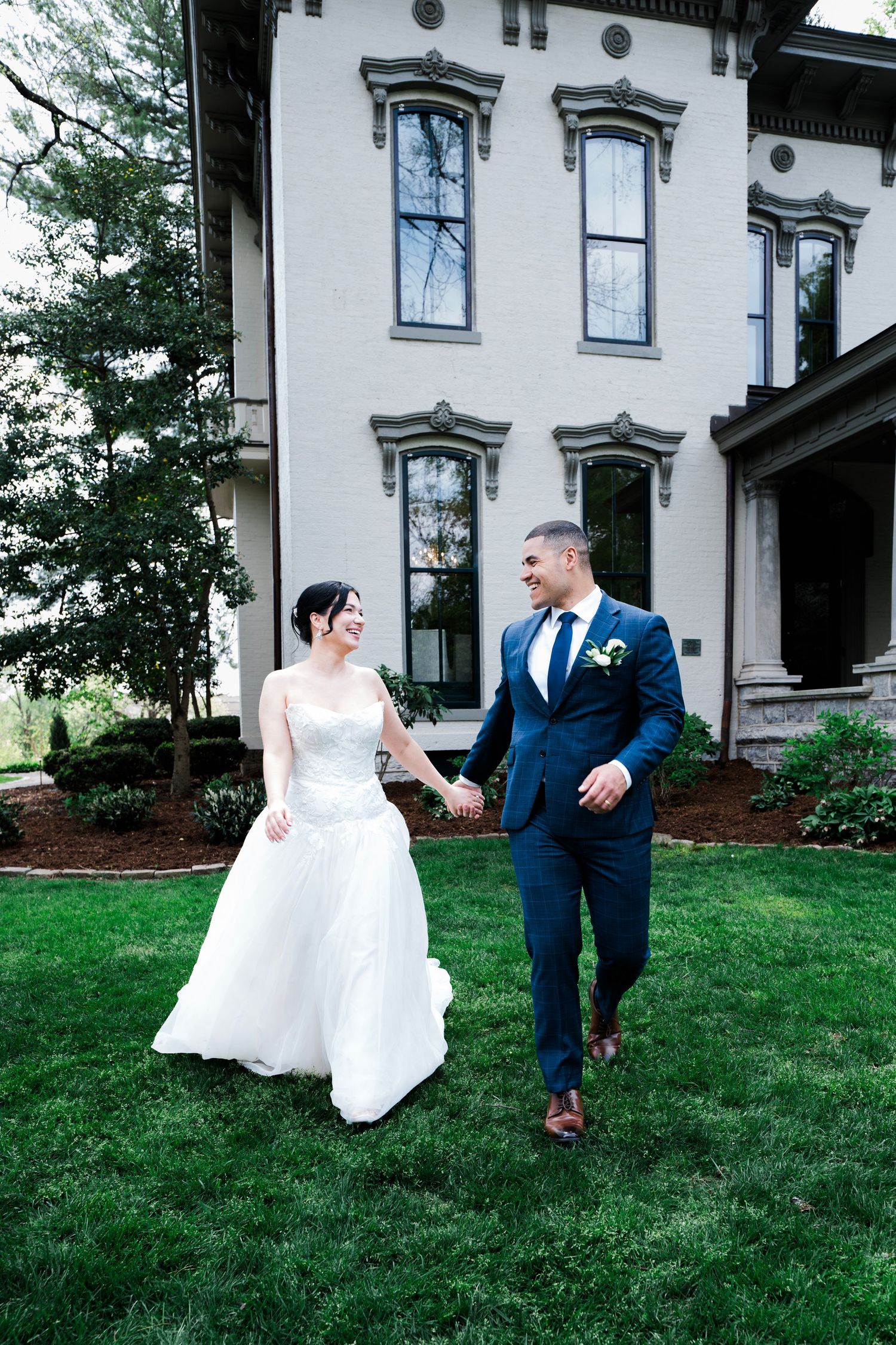 A bride and groom hold hands walking on green grass in front of an elegant historic mansion on their wedding day.