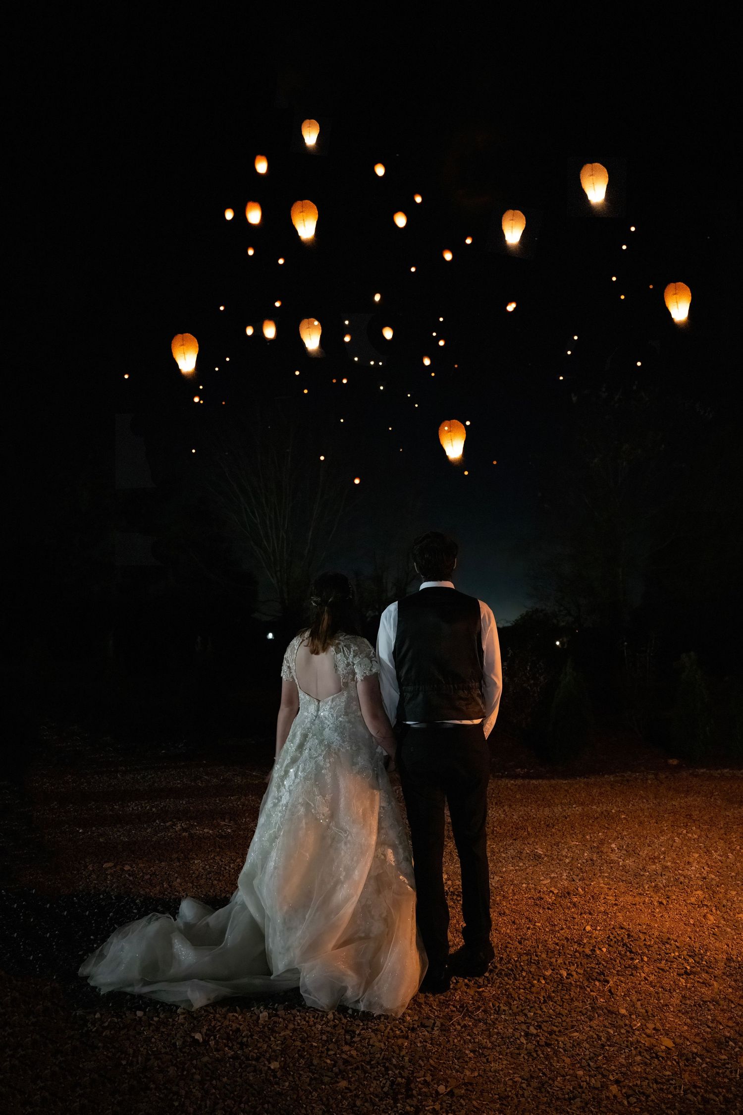 A bride and groom stand hand in hand watching glowing sky lanterns float into the dark night sky.