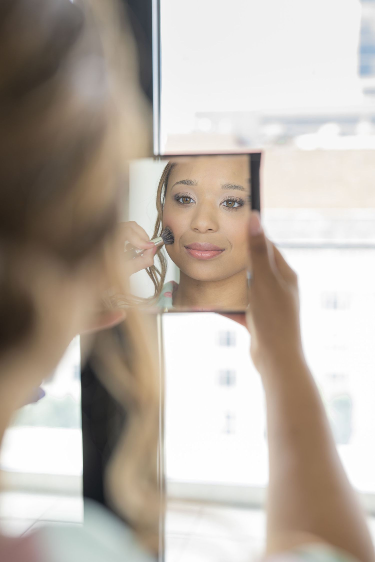 Young woman applying makeup while looking at her reflection in a handheld mirror near a bright window.