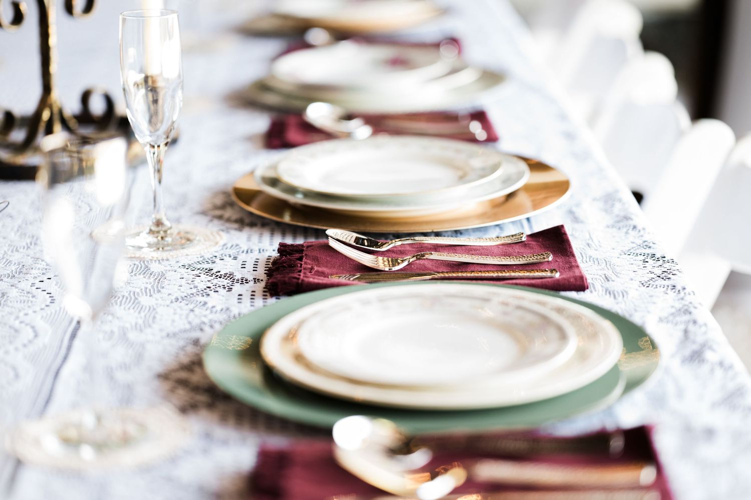 Elegant table setting with gold-rimmed plates, silver cutlery, and burgundy napkins on a white lace tablecloth.