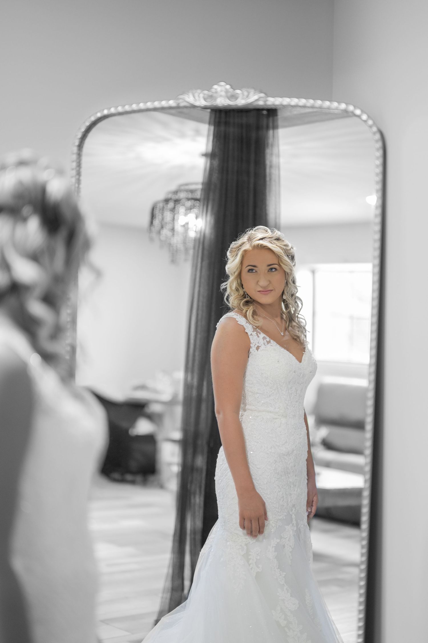 Bride in lace mermaid wedding dress gazes at her reflection in an ornate full-length mirror in a bright room.