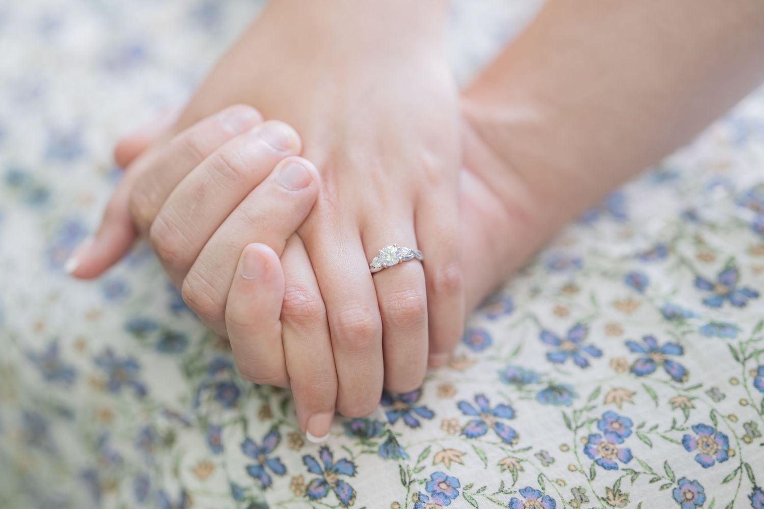 Close-up of two hands resting together, one adorned with a sparkling diamond engagement ring on a floral fabric.