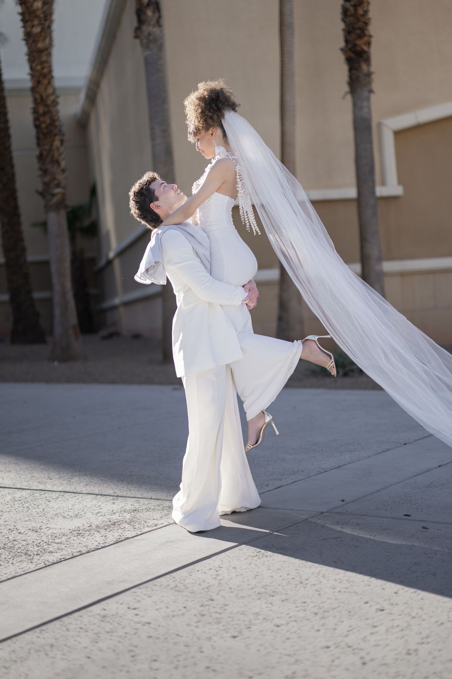 Groom in white suit lifts bride in flowing veil outdoors near palm trees on their wedding day.