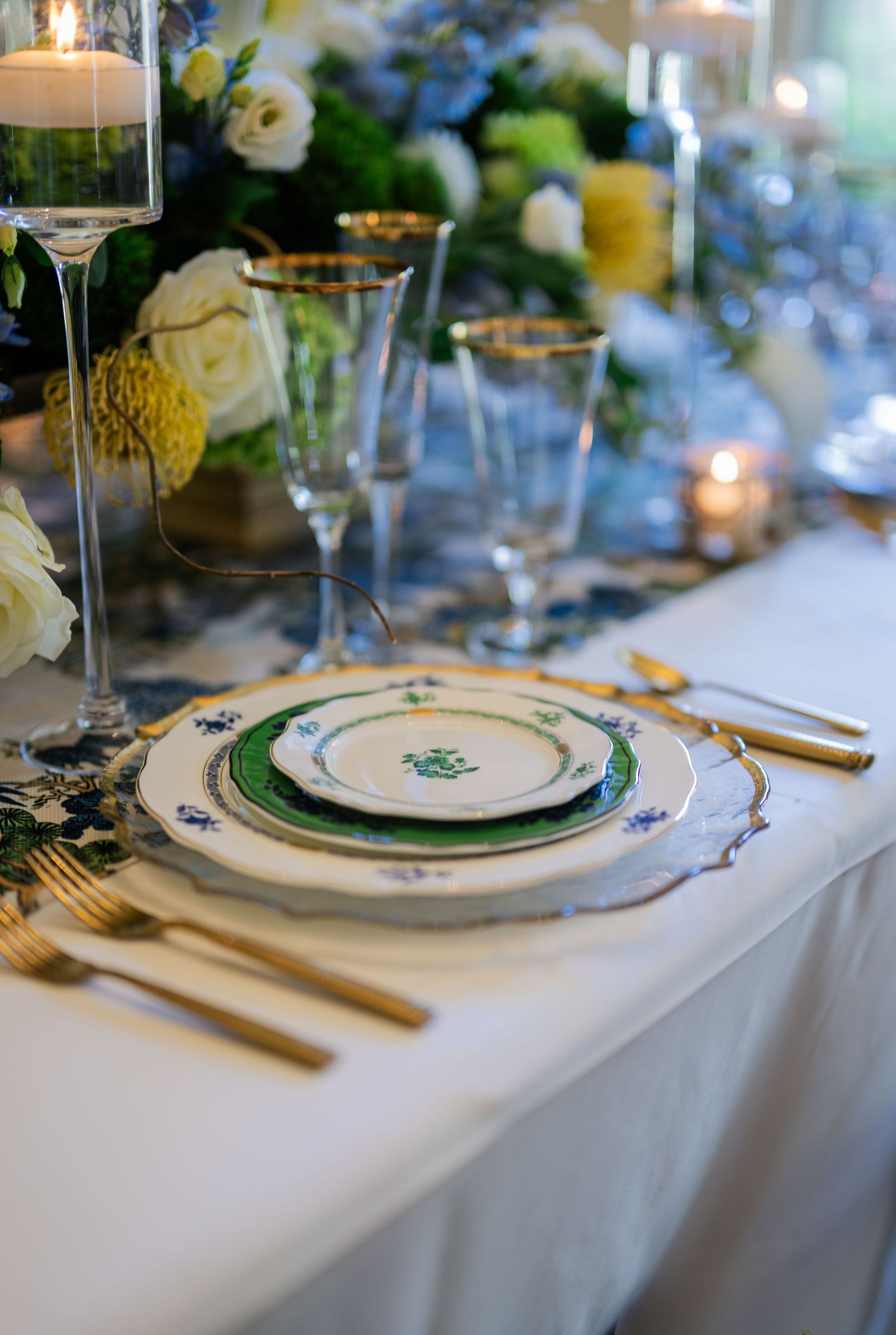 Elegant wedding table setting with gold flatware, blue floral china, crystal glasses, and white floral centerpiece.