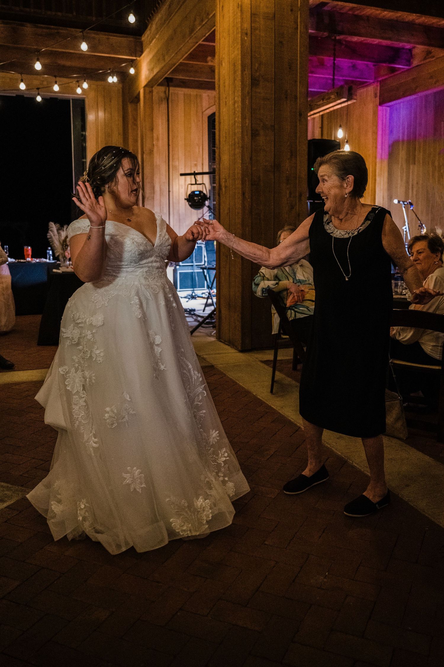 Bride in white floral gown dancing with elderly woman in black dress at rustic barn wedding reception.