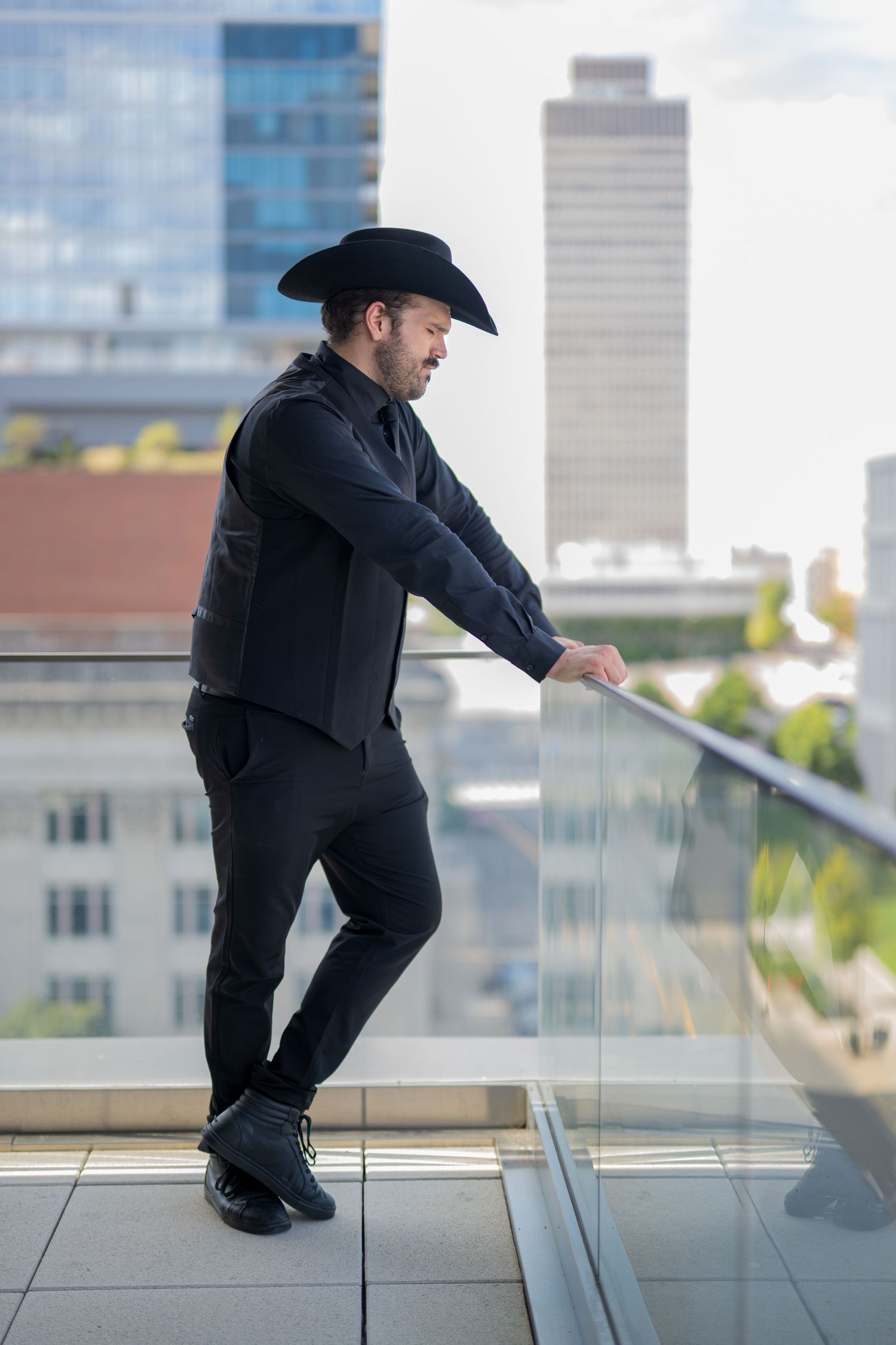 Man in black cowboy hat and all-black outfit leaning on glass balcony railing with city skyline background.