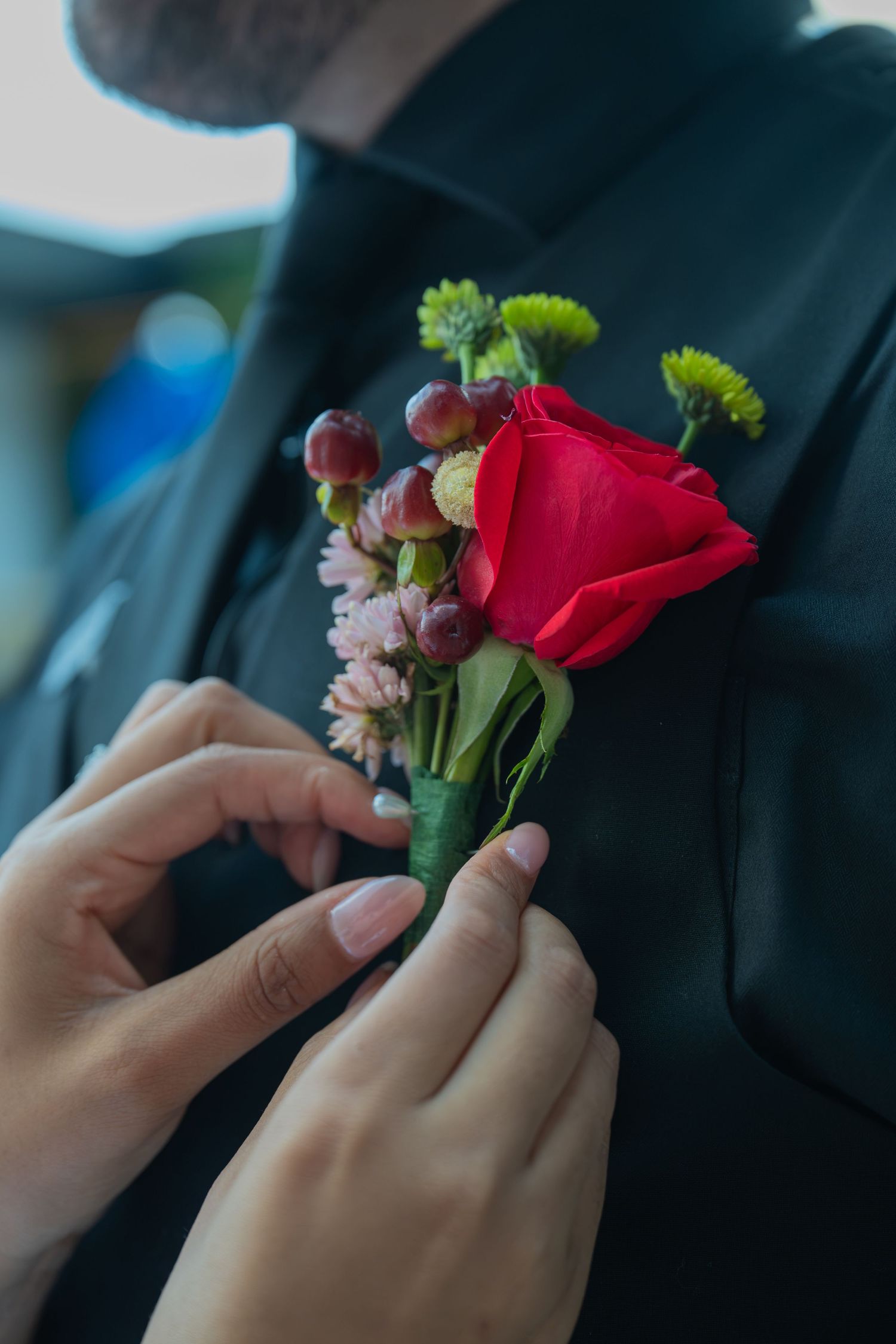 Hands pinning a red rose boutonniere with pink flowers and berries onto a dark suit jacket lapel.