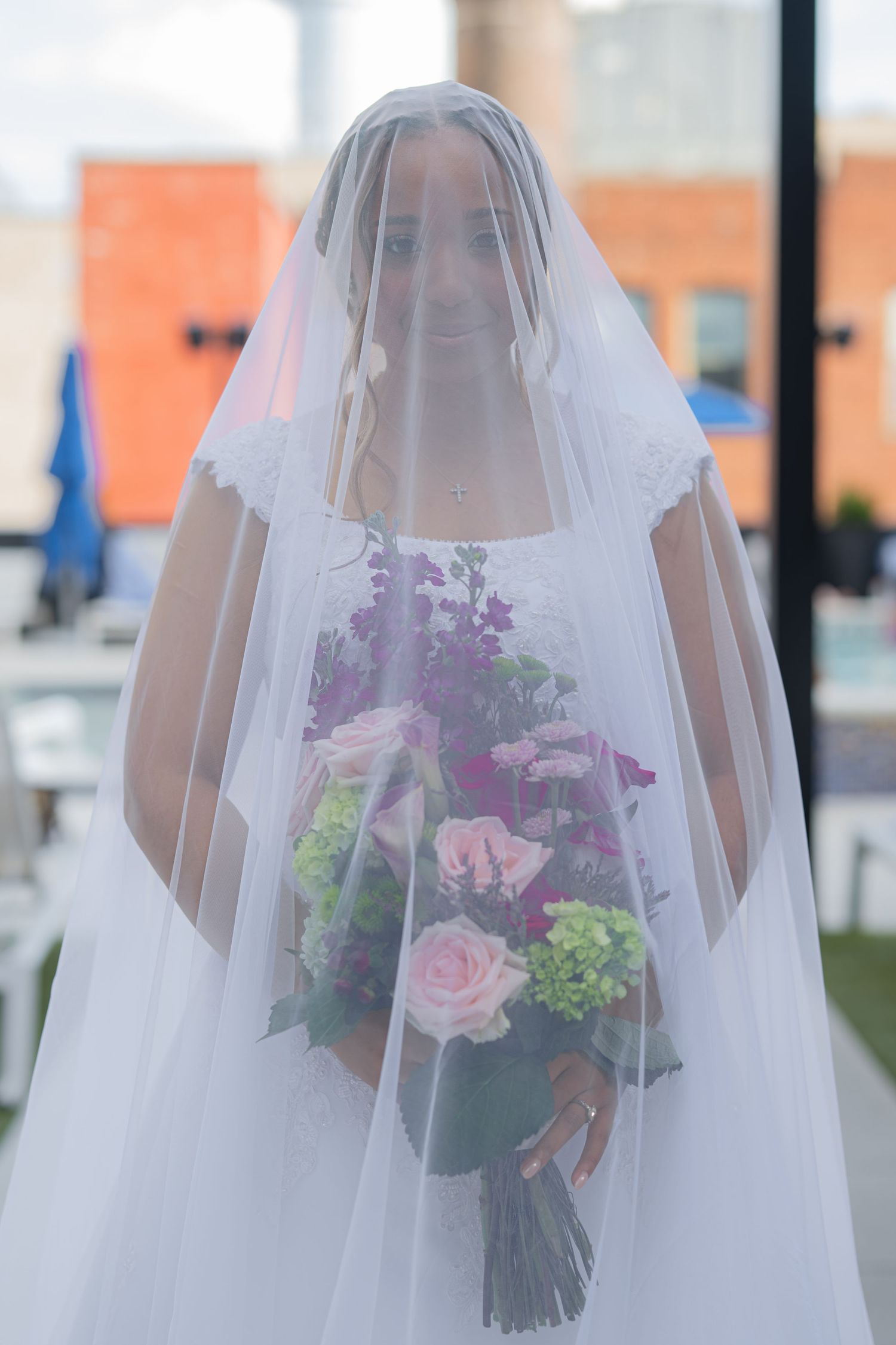 Bride in white gown and veil holding colorful bouquet of pink roses and purple flowers outdoors.