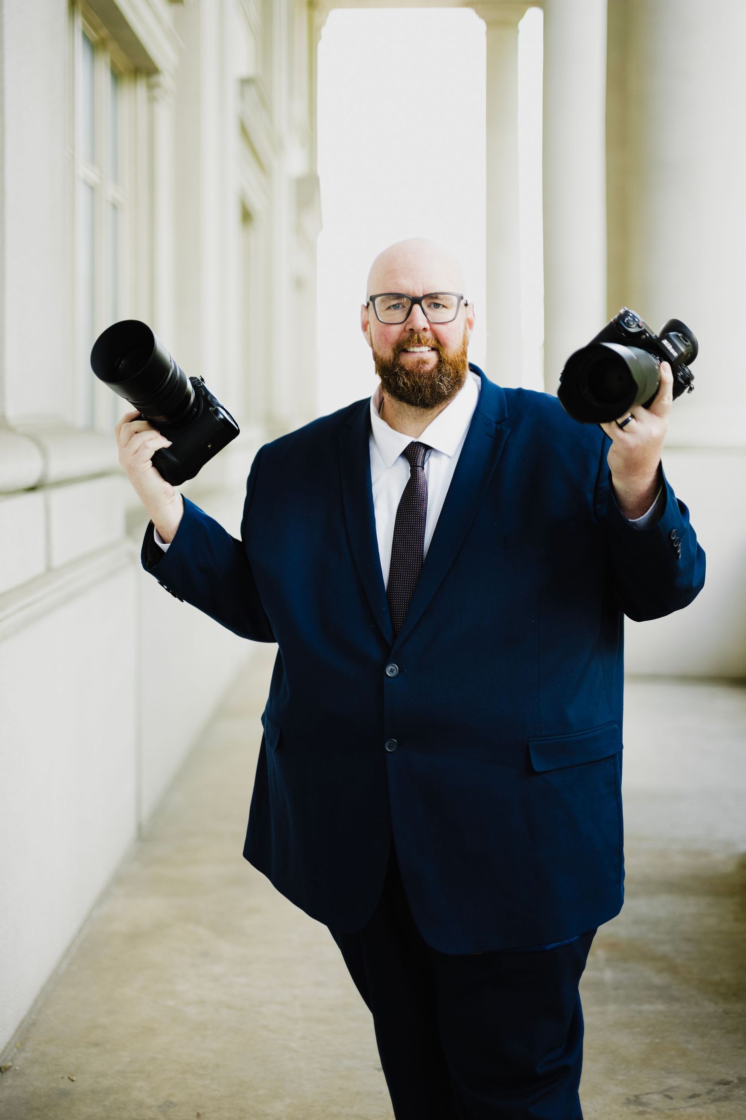 Bald bearded man in navy suit holding two professional cameras in a columned hallway.