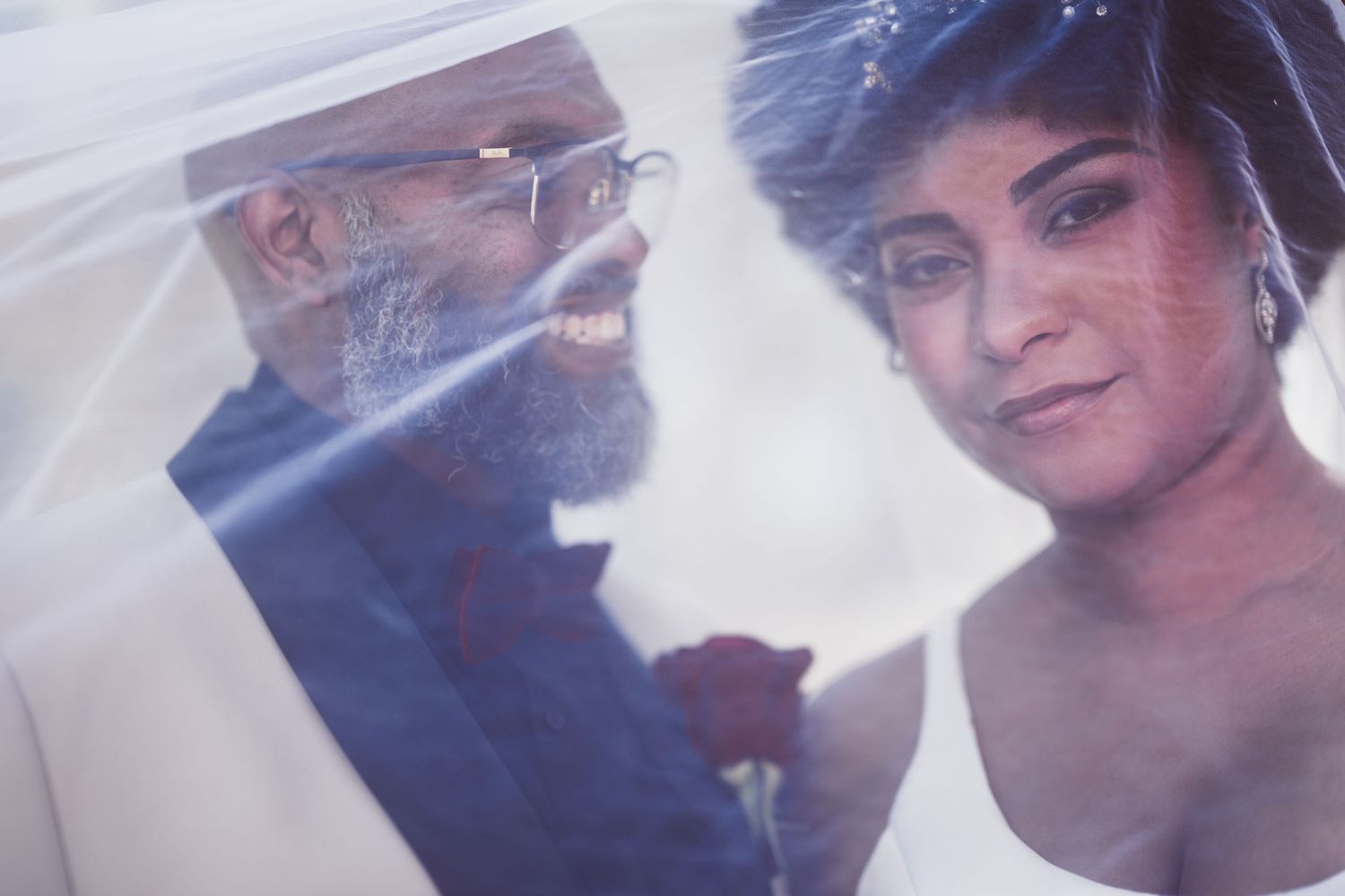 A bride and groom share an intimate moment under a flowing veil, the groom with a gray beard holding a red rose.