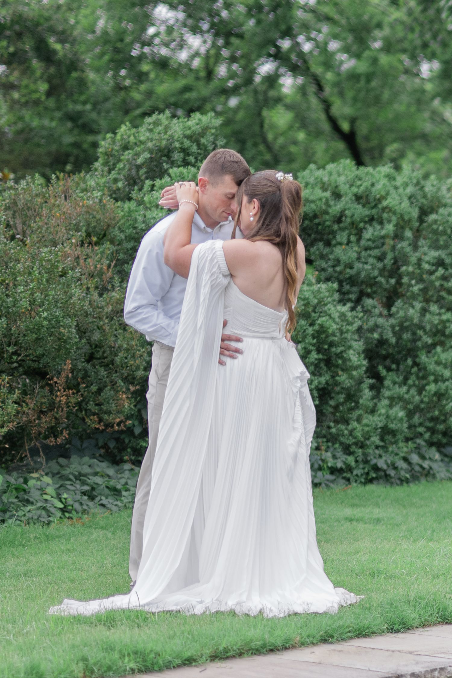 Bride and groom sharing a romantic kiss outdoors in a lush green garden, bride wearing an elegant white flowing gown.