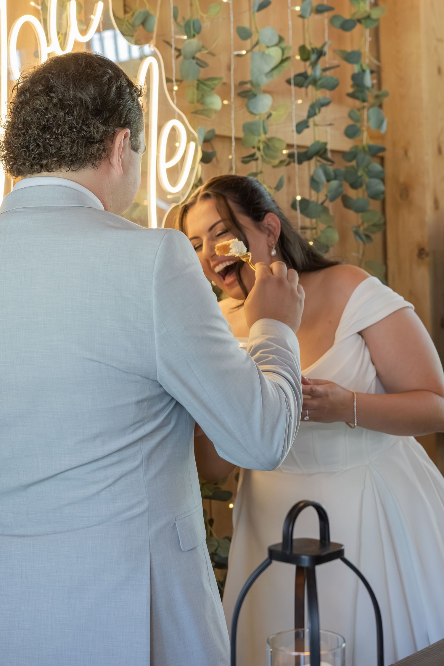 Groom feeding wedding cake to bride at reception with neon sign and eucalyptus decor in background.