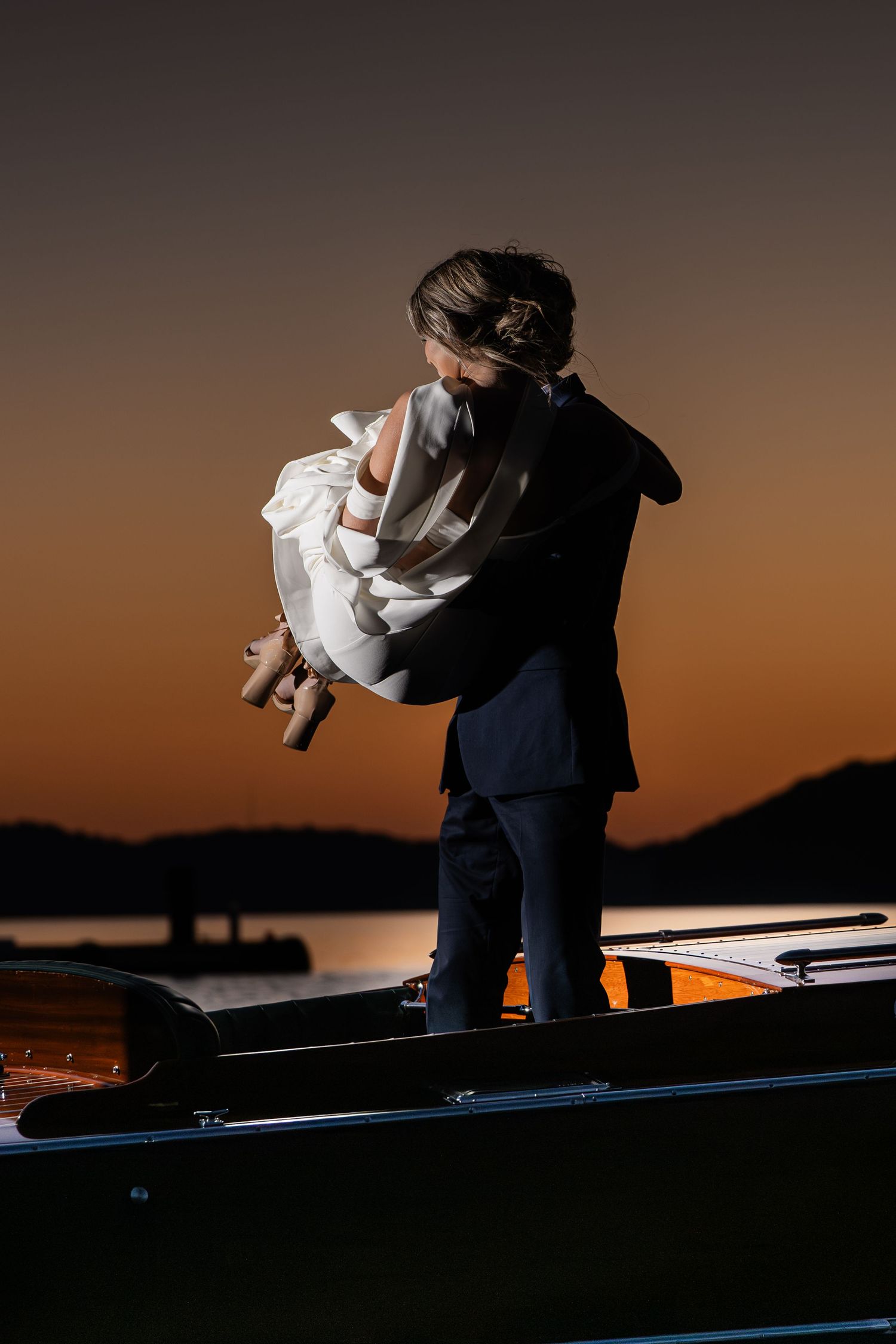 Couple embracing on a boat at sunset, silhouetted against a warm orange sky over calm water.