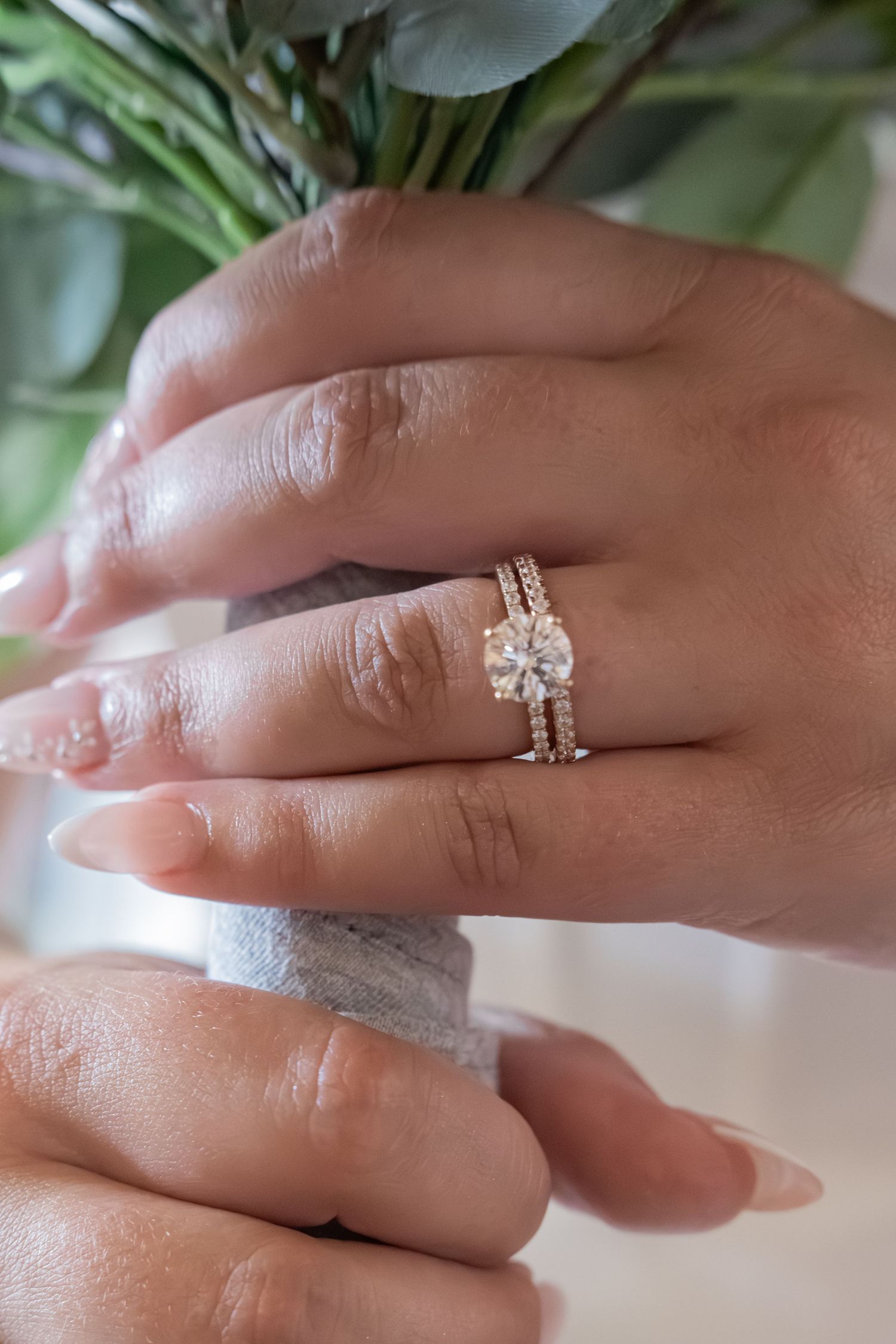 Close-up of hands wearing a stunning round diamond engagement ring with a diamond-studded gold wedding band, with greenery in background.
