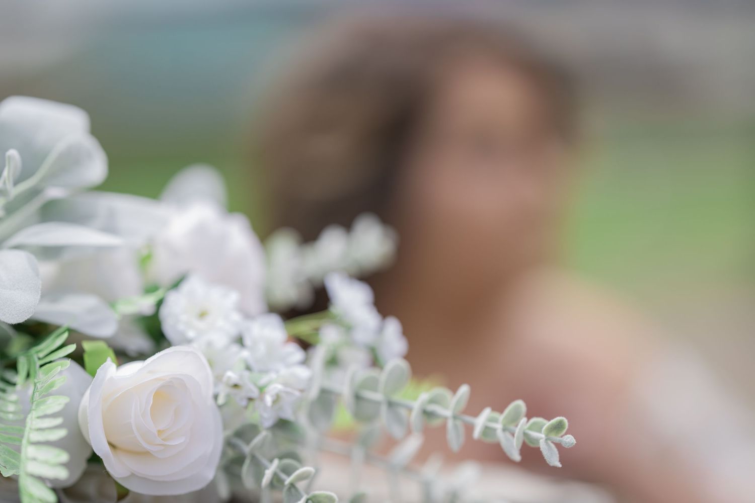 Close-up of white roses and eucalyptus bridal bouquet with blurred bride in background.