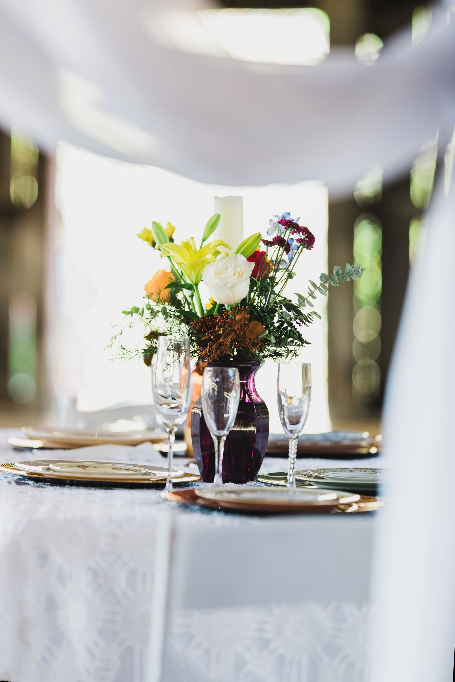 Elegant wedding table setting with colorful floral centerpiece, crystal wine glasses, and gold charger plates under white draping.