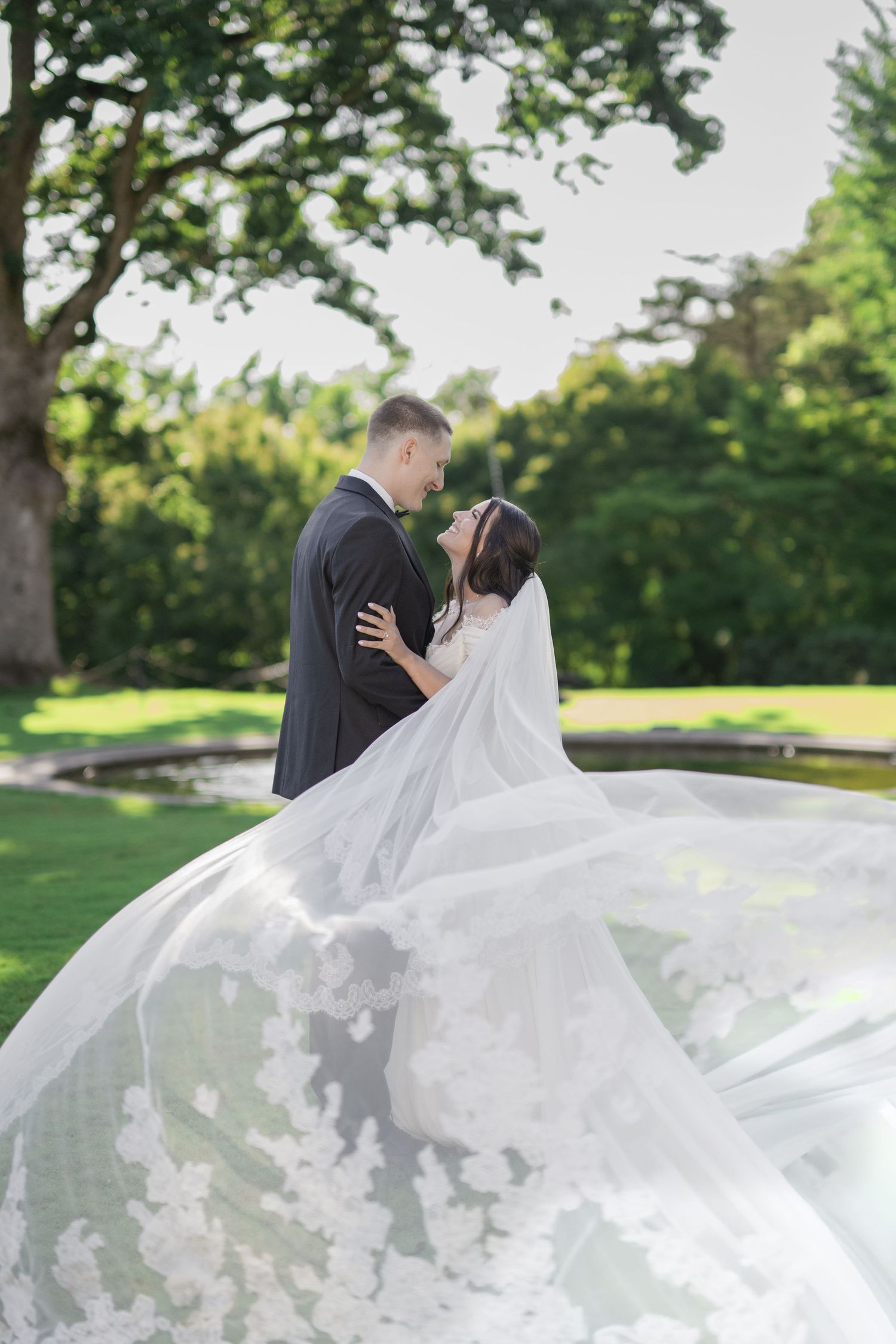 Bride and groom share a romantic kiss outdoors, with her floral lace veil flowing beautifully in the breeze.