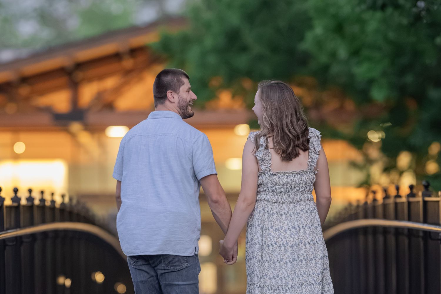 A couple holds hands while walking on a bridge at dusk, with warm lights and lush greenery in the background.