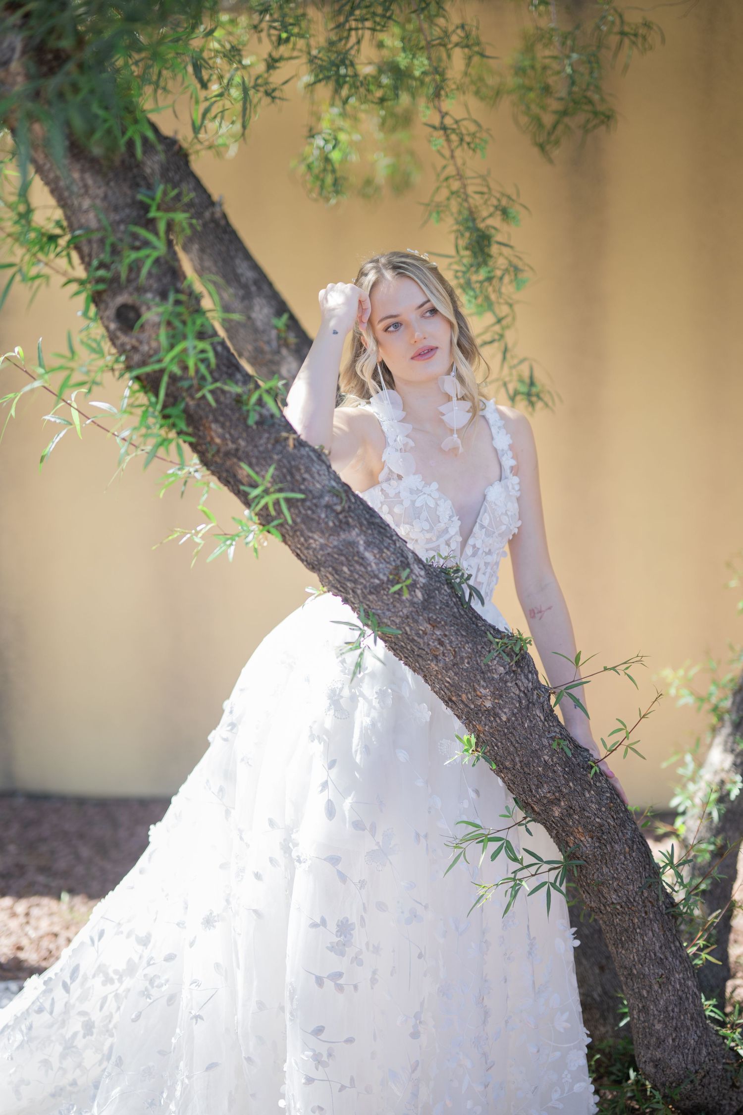 Bride in floral appliqué ball gown leans against a tree with green leaves in a sunlit outdoor setting.