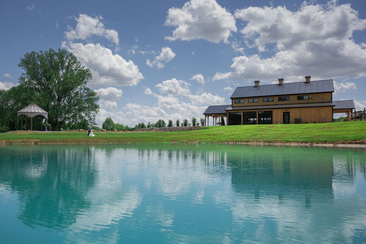 Scenic farm estate with turquoise pond reflecting clouds, green lawns, gazebo, and large barn building on a sunny day.