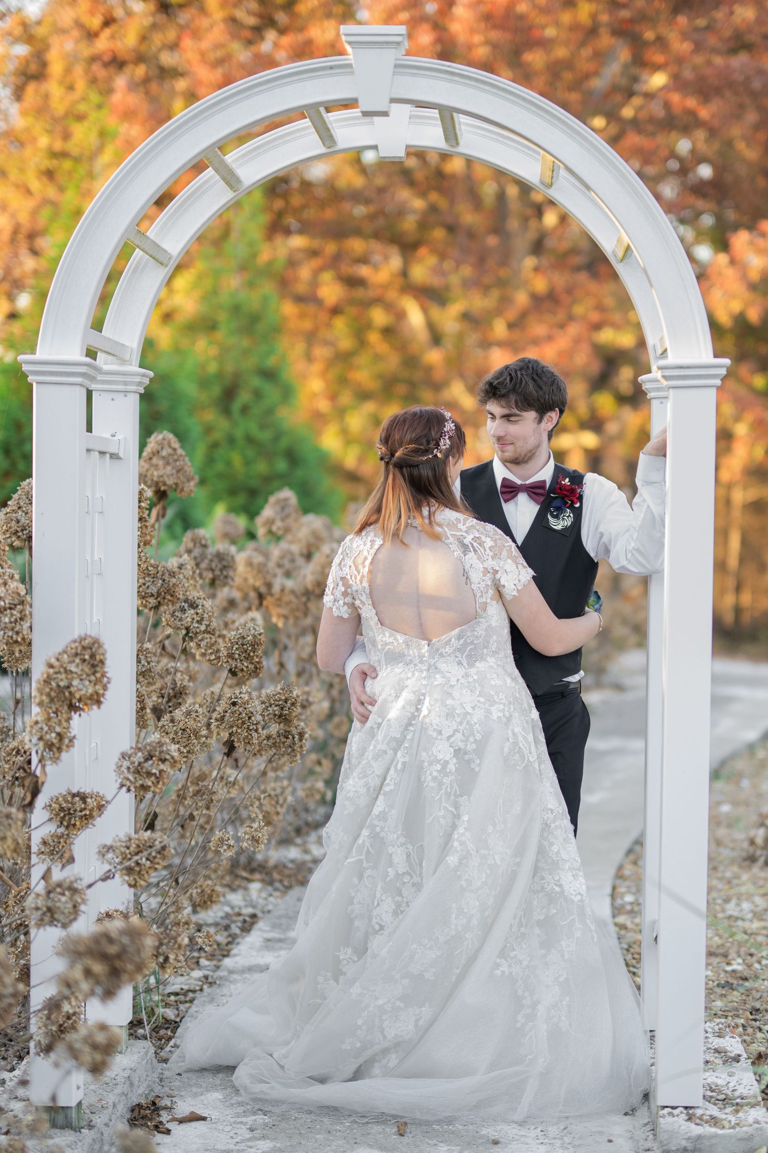 Bride and groom dancing under white arch with autumn foliage backdrop in elegant wedding attire.