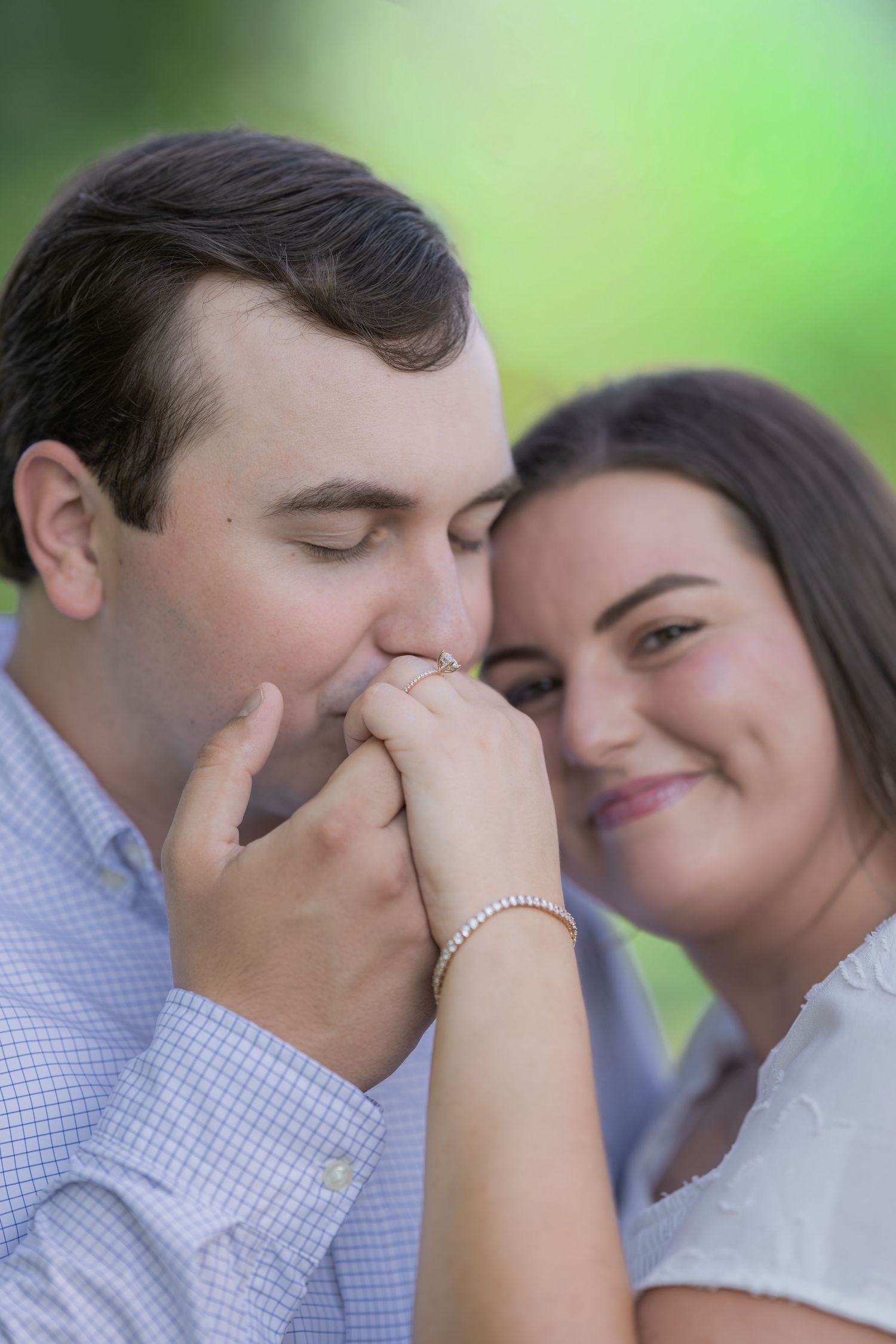 A man tenderly kisses a woman's hand as she smiles warmly, showcasing an engagement ring outdoors.