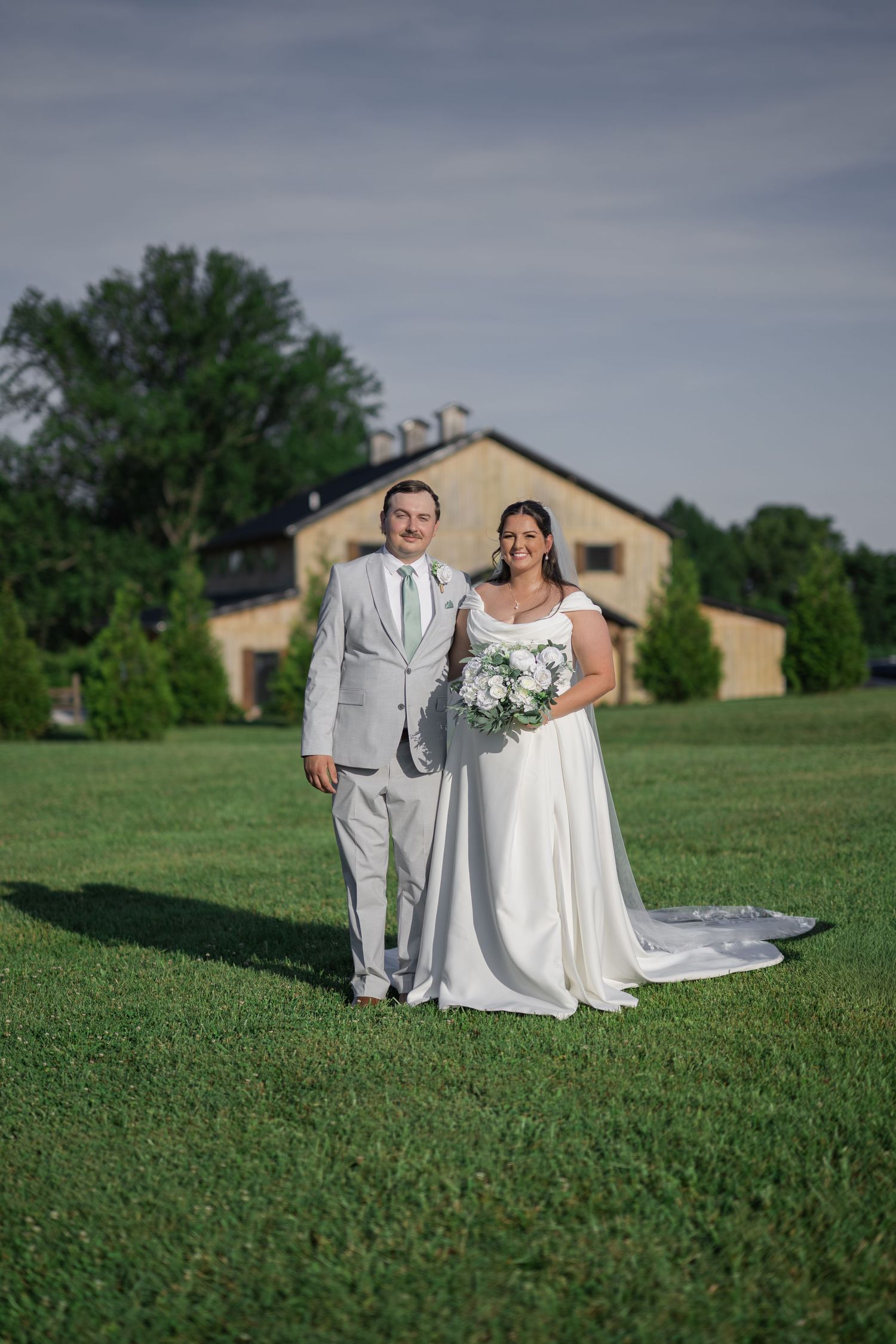 Bride and groom posing on green lawn with rustic barn venue behind them on their wedding day.