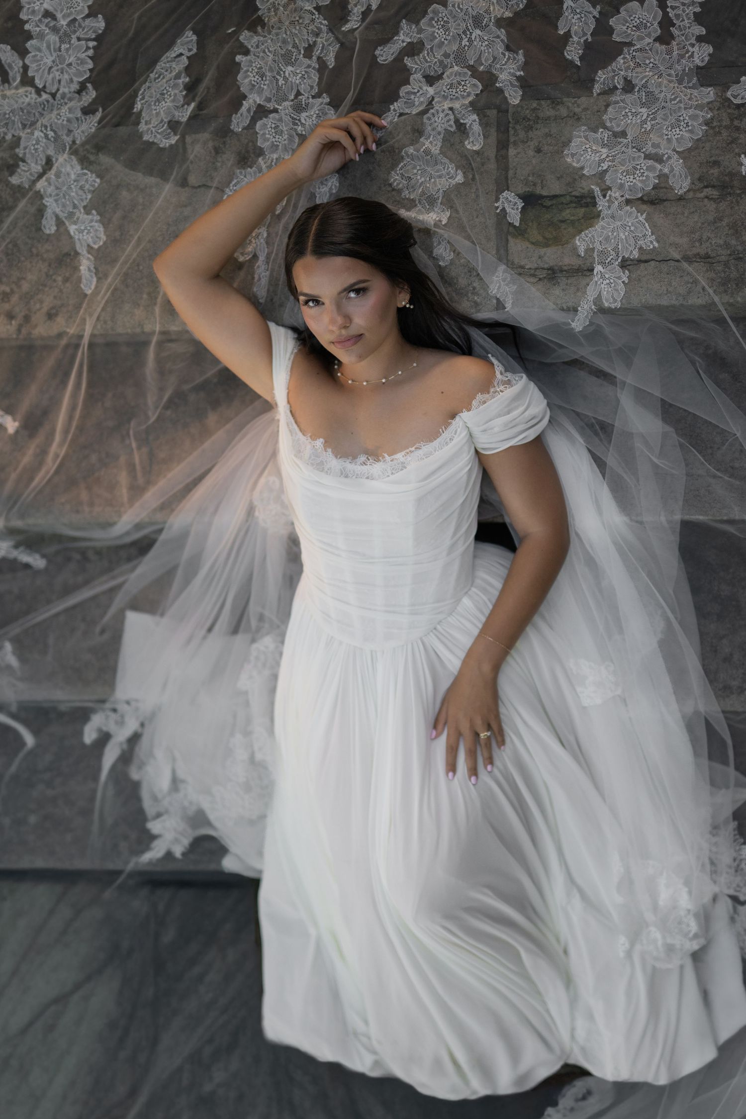 Bride in white chiffon off-shoulder wedding dress with lace veil backdrop, posing elegantly in a bridal portrait.