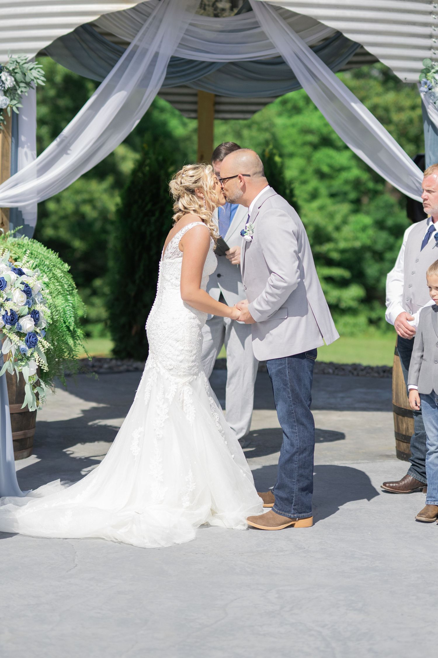 Bride and groom share first kiss at outdoor wedding ceremony beneath white draped arch with blue floral arrangements.