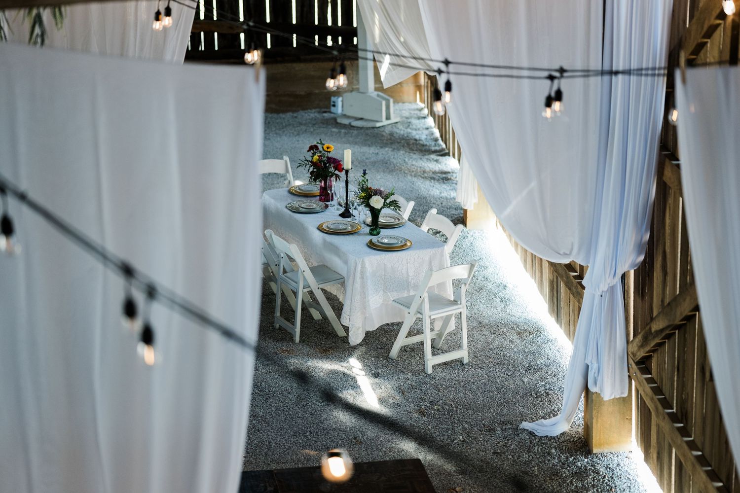 Elegant barn wedding reception with white draping, string lights, white folding chairs, and floral centerpieces on draped tables.