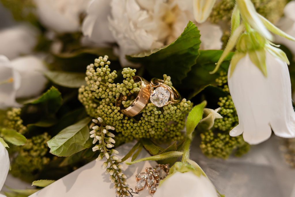 Gold wedding bands and diamond engagement ring resting on a lush bridal bouquet with white flowers and green berries.