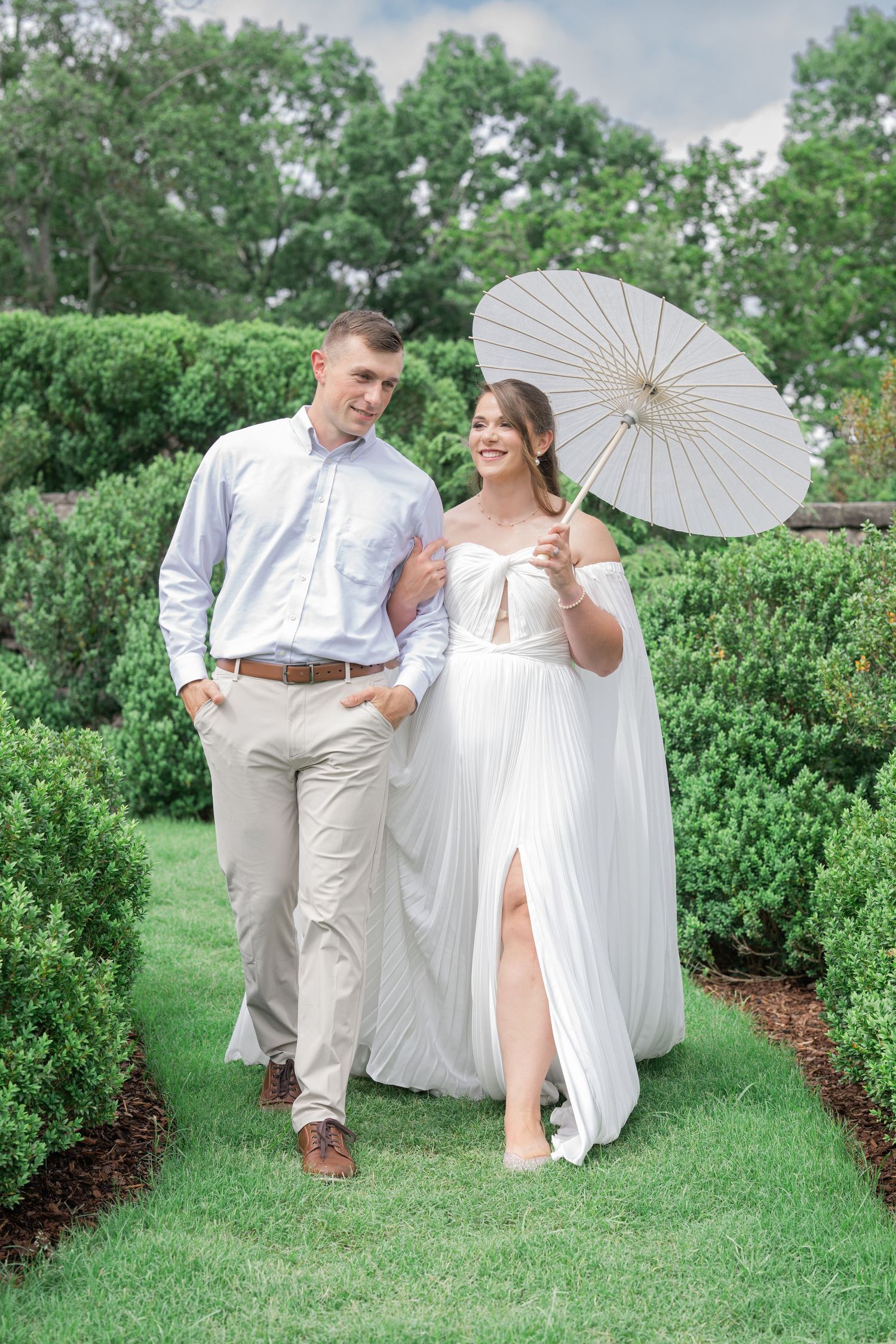 Couple walks through garden, bride in white gown holds parasol, groom in khakis, lush green hedges background.
