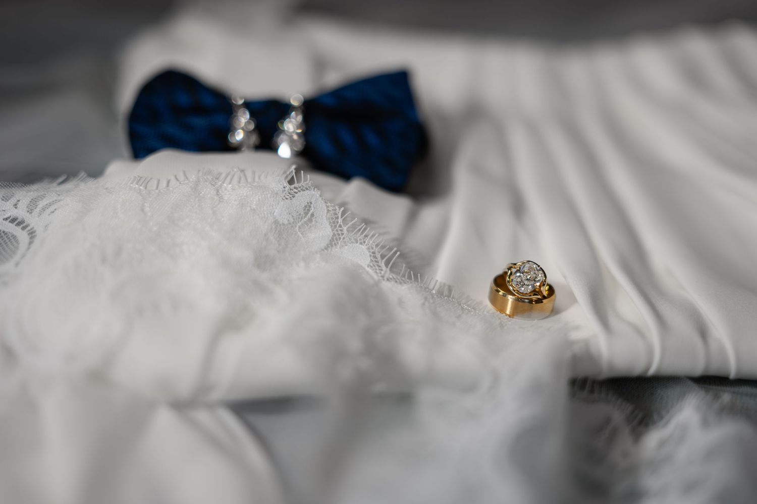 Gold diamond engagement ring and blue bow tie with pearl earrings on white lace wedding dress fabric.