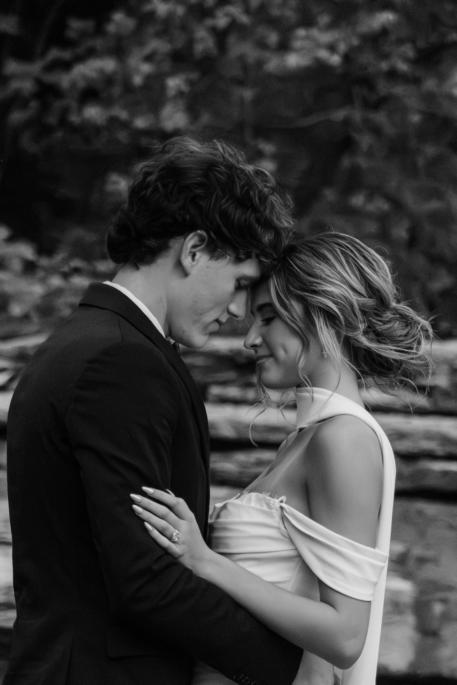 Black and white photo of a couple in wedding attire sharing an intimate moment outdoors among lush greenery.