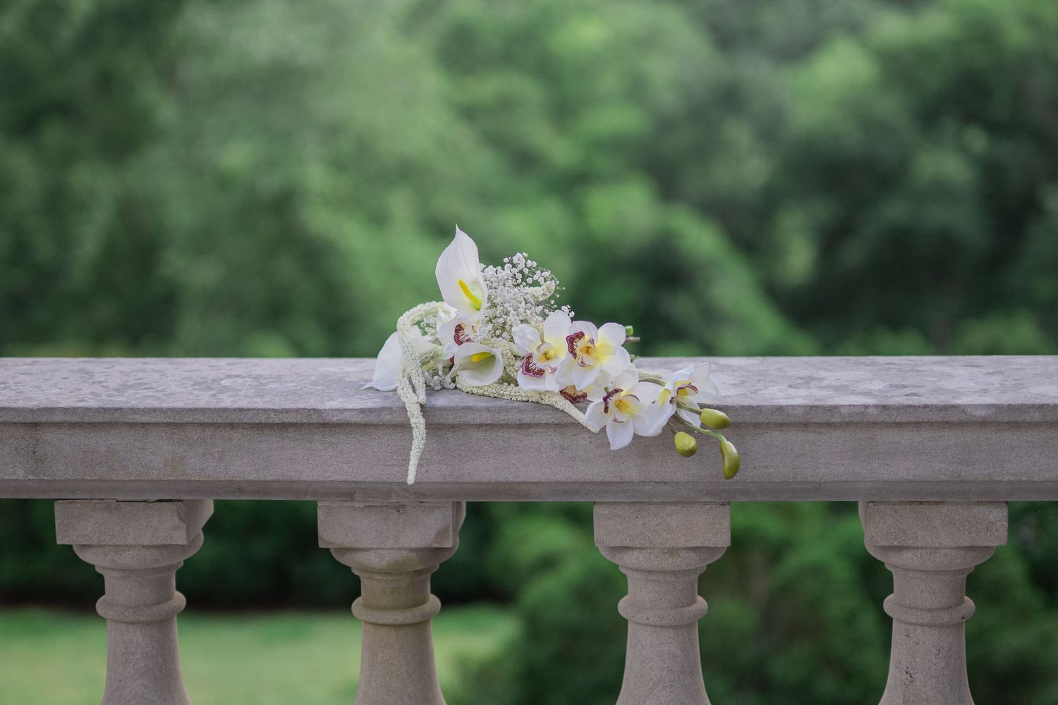 White orchid bouquet resting on a stone balustrade railing with lush green trees in the background.