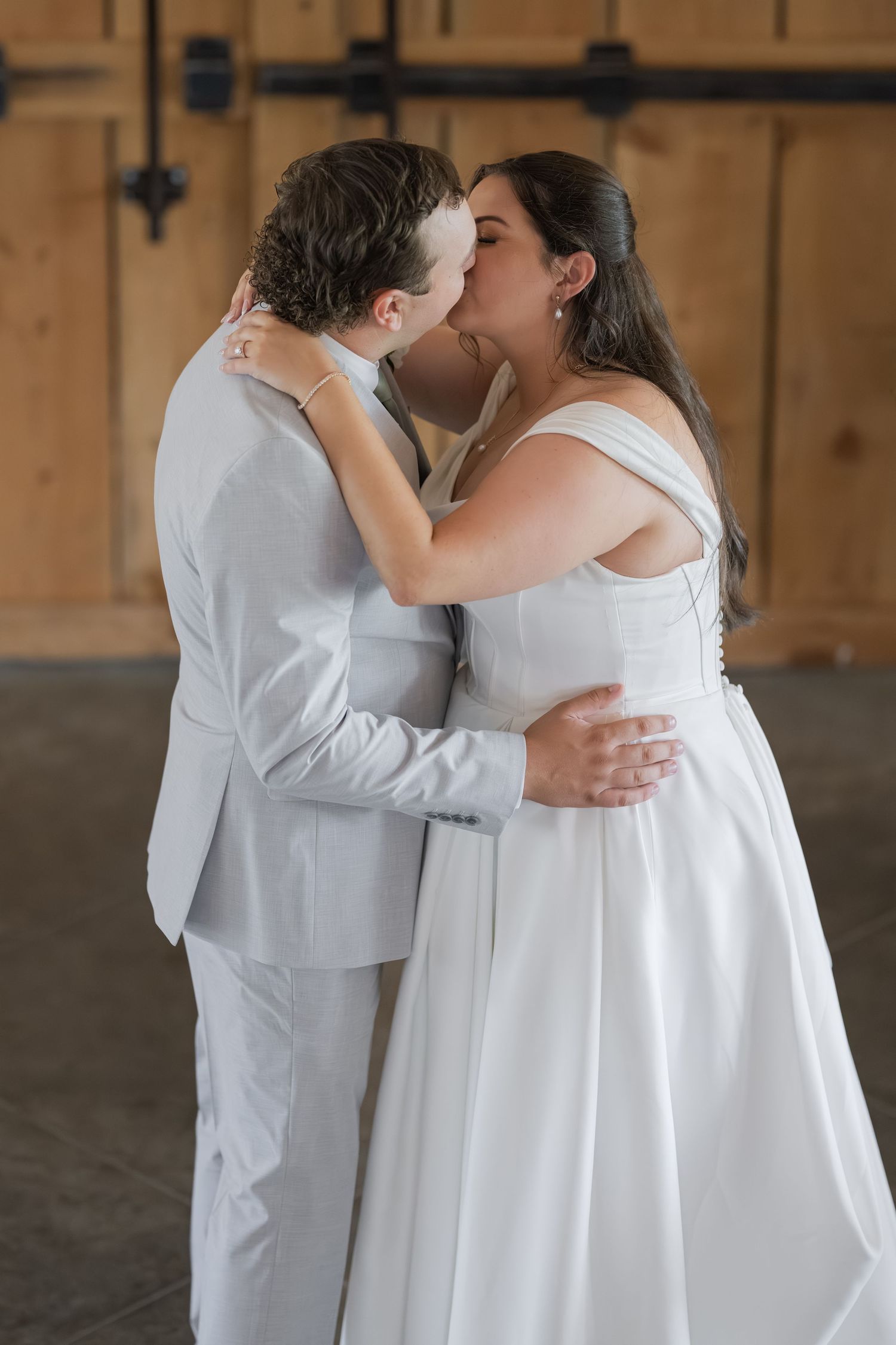 Bride and groom sharing their first kiss in a rustic barn venue, both dressed in white wedding attire.