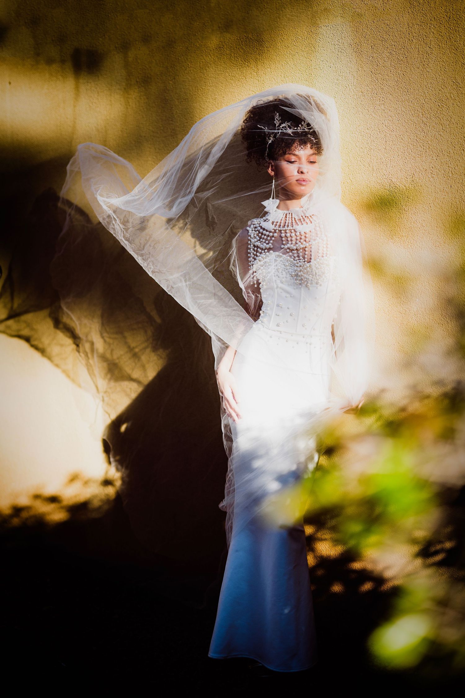 Bride in white lace gown and flowing veil stands gracefully against a sunlit wall, wearing a delicate tiara.
