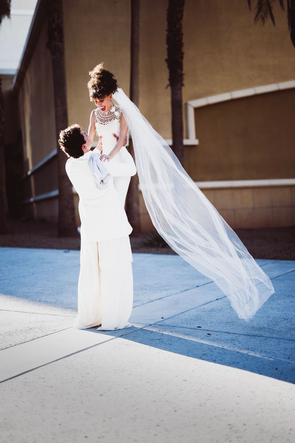 Groom lifting bride in white wedding dress and veil outdoors on sunny day.
