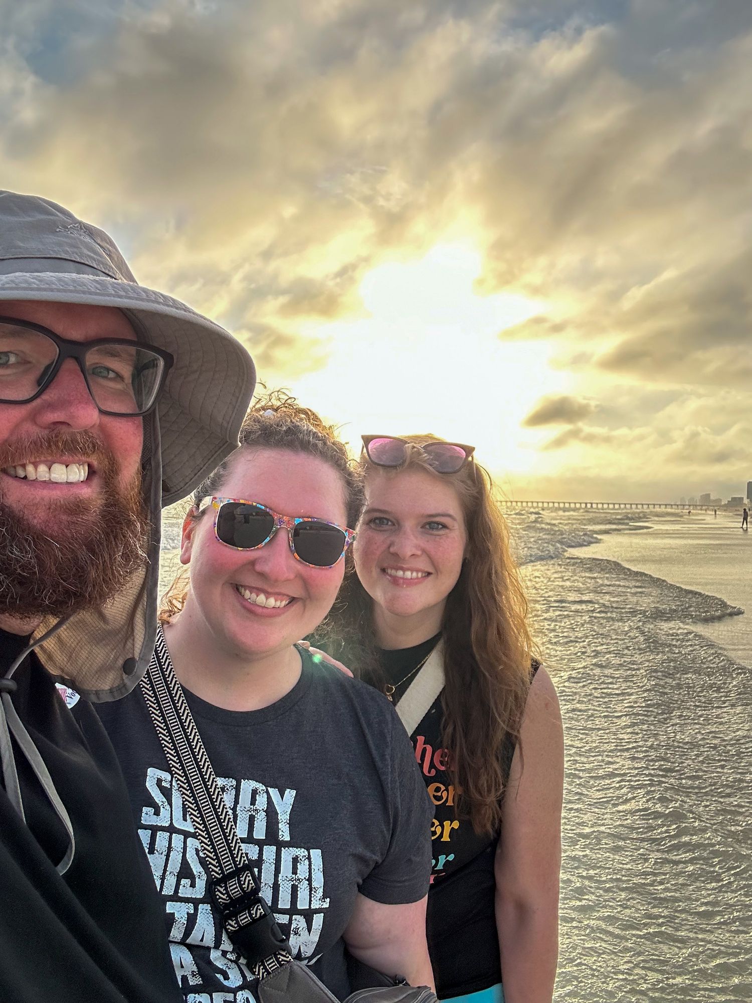 Three friends smile in a selfie at sunset over a coastal bridge with dramatic golden clouds in the background.