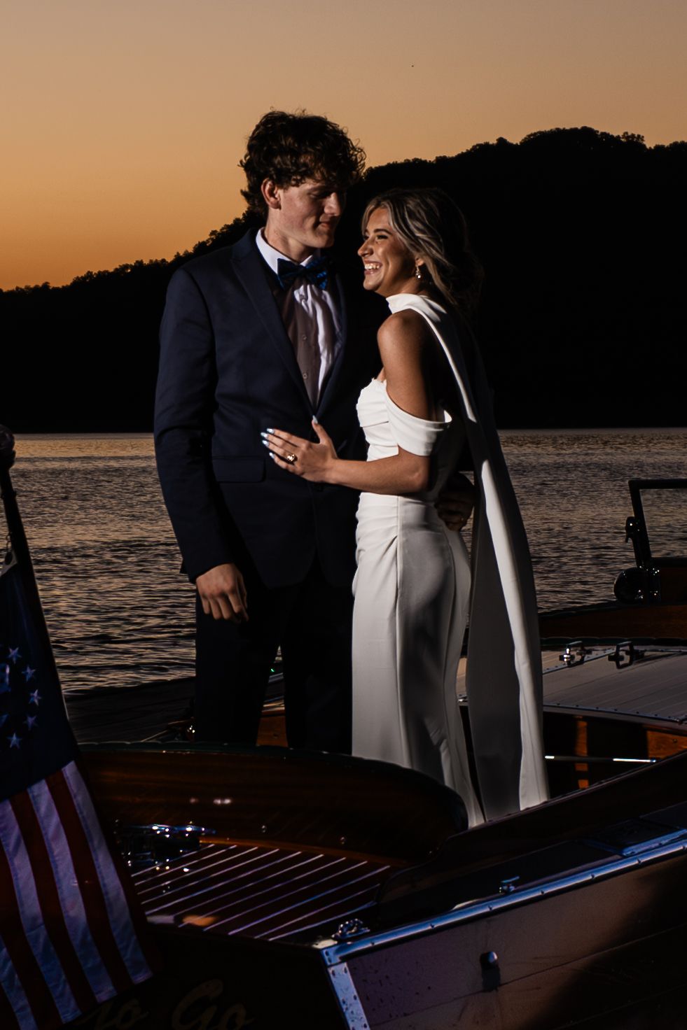 Couple in wedding attire embracing at sunset on a lakeside dock, bride in white gown, groom in navy suit.