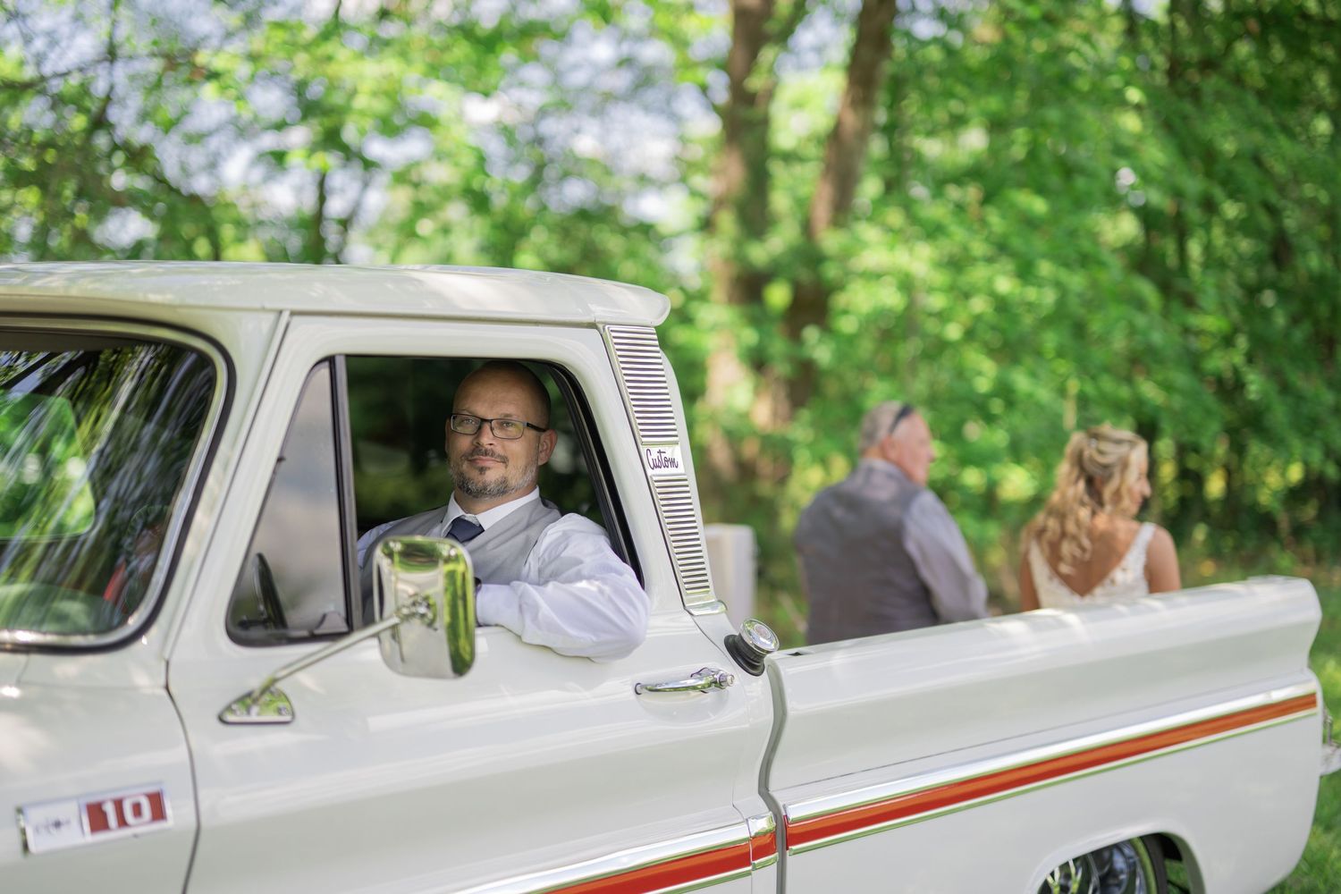 Man in white shirt smiling from vintage white truck at outdoor wedding ceremony with bride visible in background.