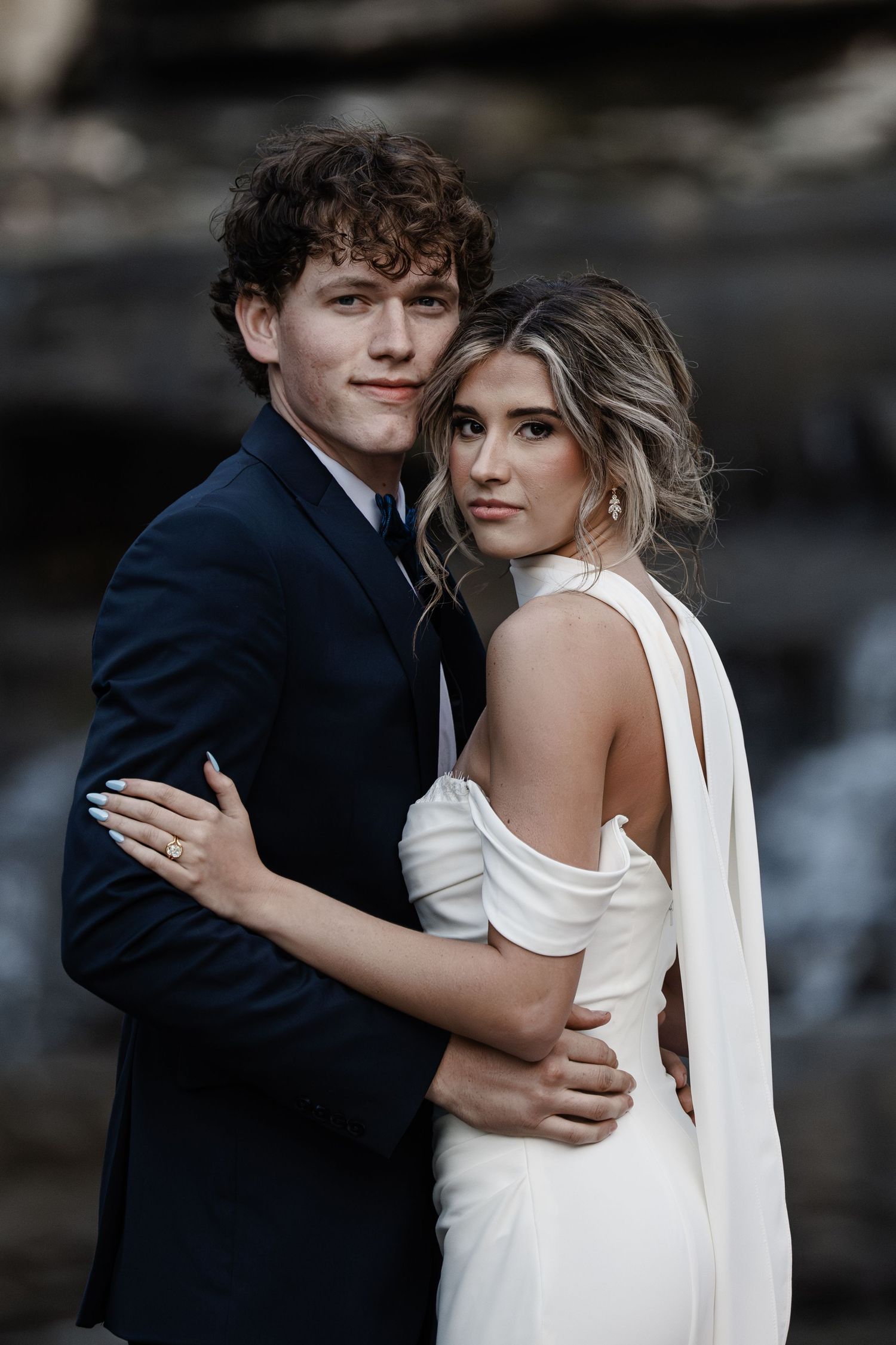 A young couple in wedding attire embrace outdoors, groom in dark suit, bride in elegant backless white gown.